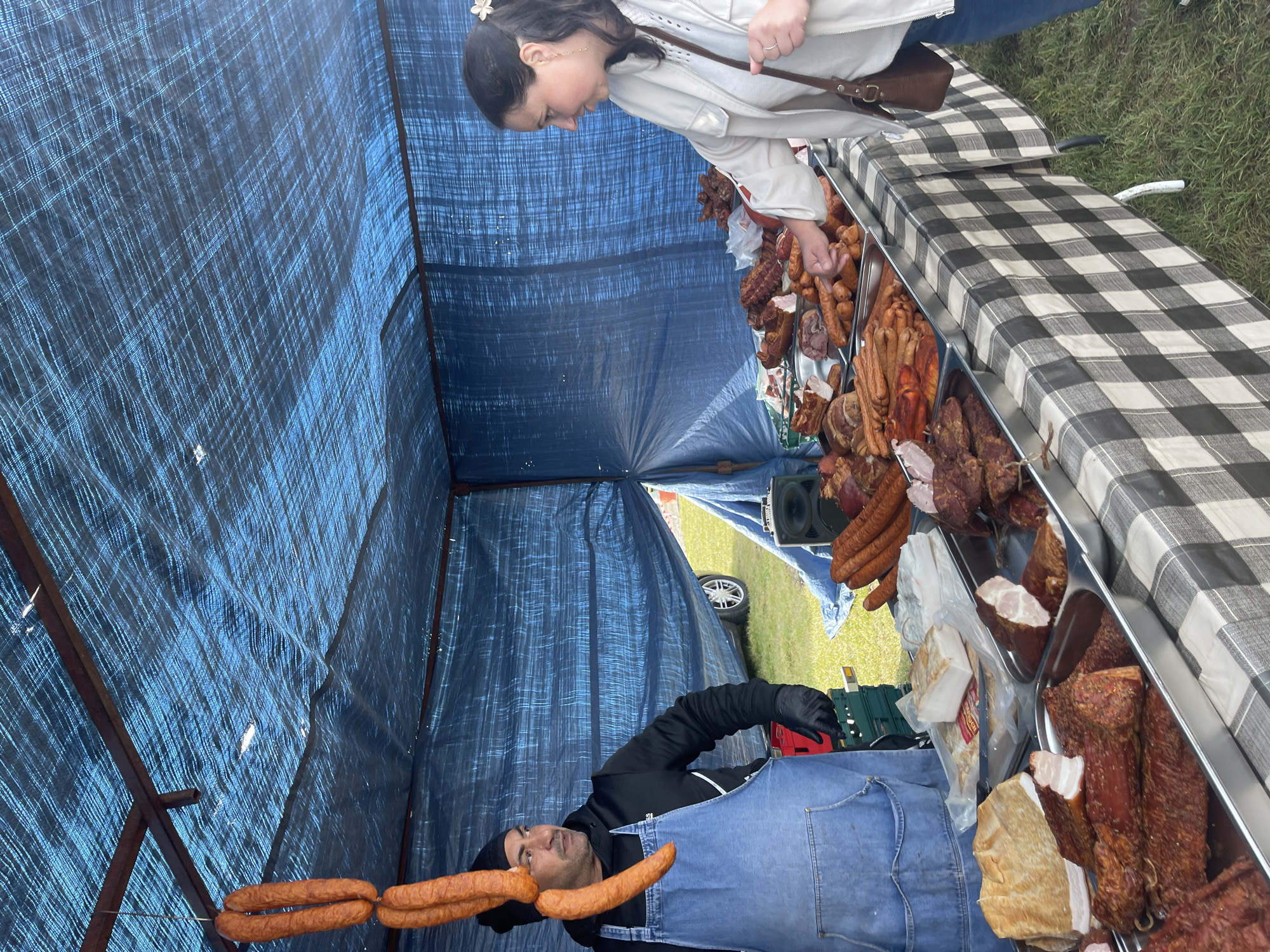 At a stall in Sevenoaks, United Kingdom, a woman in a light-colored jacket and jeans inspects sausages and cured meats displayed in metal trays on a checkered tablecloth. A vendor wearing a black hooded jacket and a denim apron stands behind the counter. Sausages hang overhead. The stall is covered with a blue tarp. Grass is visible in the background.