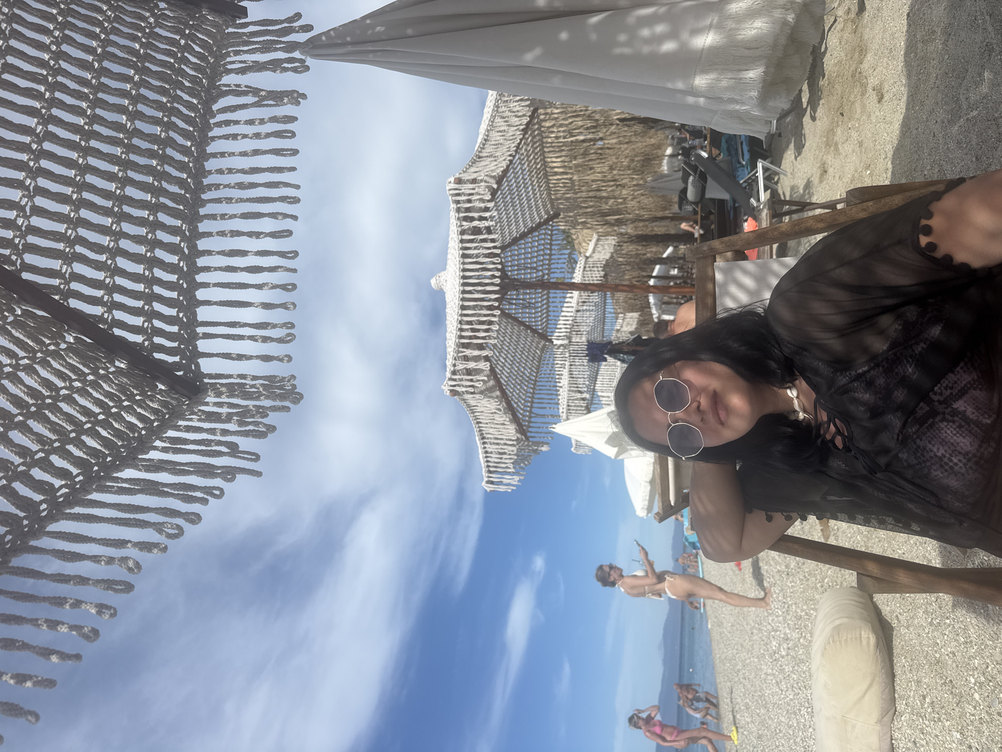 The image shows a young woman sitting in a beach chair, facing the camera. She is wearing sunglasses and a sheer black cover-up over a patterned garment. She has a shell necklace on. Behind her, the beach is populated with other beachgoers, beach umbrellas, and loungers. The ocean is visible in the background, under a partly cloudy blue sky. The scene appears to be in Pallini Municipal Unit, Greece, likely a beach resort area.