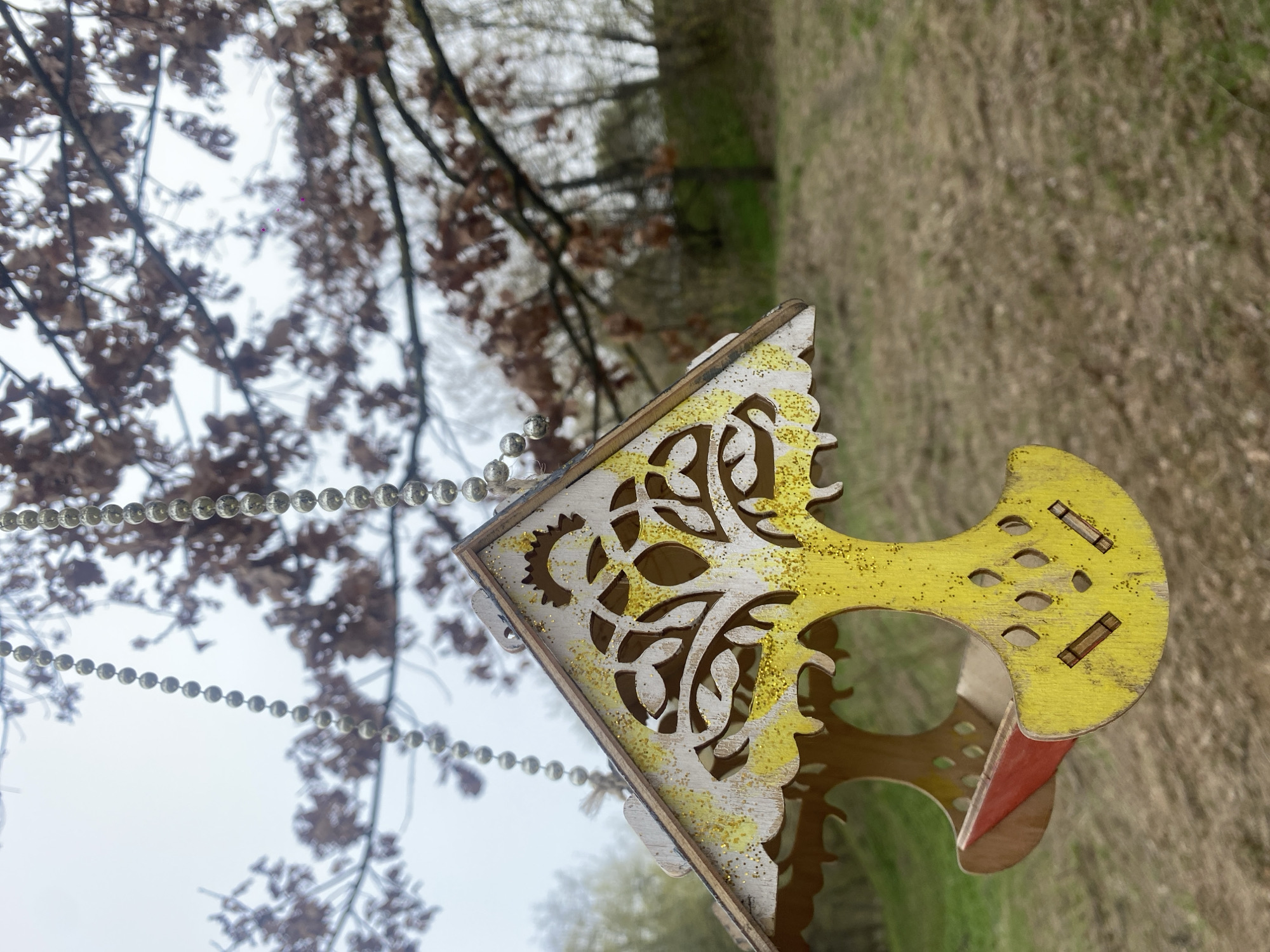 This media file captures a close-up of a decorative wooden ornament, rotated 90 degrees clockwise from its natural orientation, set against an outdoor backdrop in Moscow, Russia. The central object is crafted from wood, predominantly painted in a vibrant yellow and white with sparkling gold glitter. It features elaborate cutouts resembling intertwined foliage or ornate patterns. A small red-painted wooden piece is visible on its underside, possibly part of its base or interior. The ornament appears to be suspended by multiple strands of silver-colored, spherical beads, which hang diagonally and vertically across the upper left portion of the frame.
The background consists of a bright, overcast sky on the left, visible through the bare branches of trees that still hold some dry, brown leaves. On the right side, out of focus, is an expanse of dry, light brown ground cover, possibly grass or fallen leaves. This natural setting, with its leafless trees and dry ground, suggests the season is late autumn, winter, or early spring. There are no people visible, and no specific activity is taking place other than the object hanging, likely as a decoration. The time of day is daytime, under a cloudy sky. No text is discernible anywhere in the image.