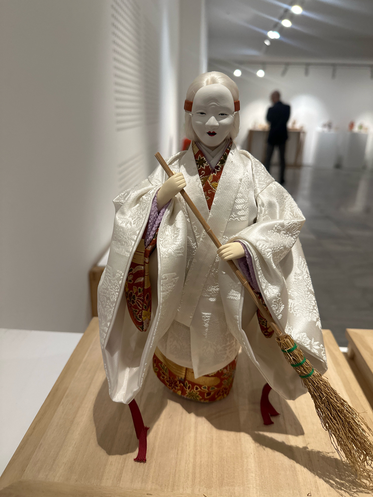 A traditional Japanese doll, possibly a Kabuki actor representation, stands on a light-colored wooden surface in what appears to be an art gallery or exhibition space. The doll wears an elaborate white kimono with intricate patterns and a colorful obi. It has a pale, mask-like face with stylized features and holds a straw broom. In the background, the blurred figures of several people are visible, suggesting this is a public space. The lighting indicates an indoor setting with overhead lights. The doll is positioned in the foreground, with the rest of the scene receding into a soft focus.