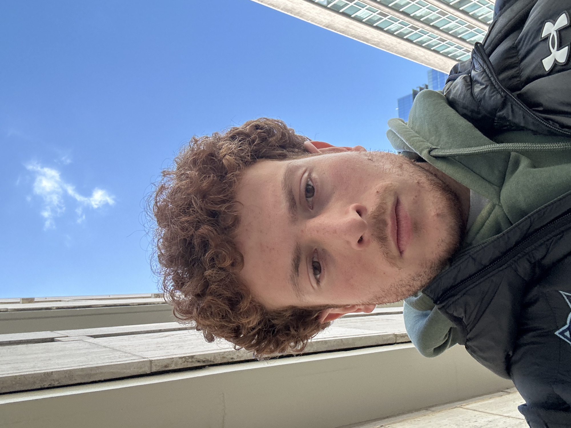 A low-angle photograph taken from above, showing a person outdoors in a city, possibly New York. The person has fair skin, short reddish-brown curly hair, and light facial stubble. The person is wearing a black puffy jacket, visible at the right edge of the frame, and a light green hooded sweatshirt beneath. The upper portion of a tall building with multiple windows is visible at the top of the frame, angled slightly. A bright blue sky with a few scattered clouds is also visible in the upper left area. In the lower part of the frame, there are architectural details of a light-colored building or structure with linear design elements.