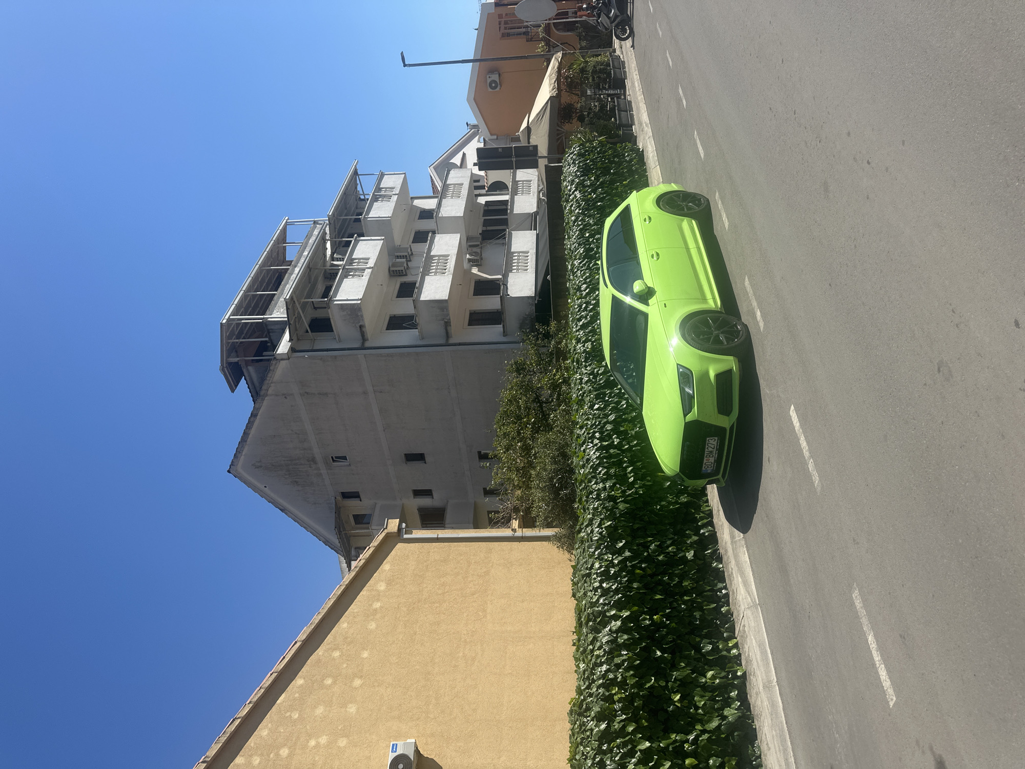 The image shows a bright green Audi parked on a street in Budva, Montenegro. The car is on the right side of the road, next to a tall hedge. Behind the hedge are two buildings, one is yellow and the other is white. The sky is clear and blue. A license plate is visible on the car, reading “BD BM 273”. The street appears to be freshly paved, with white lane markings.