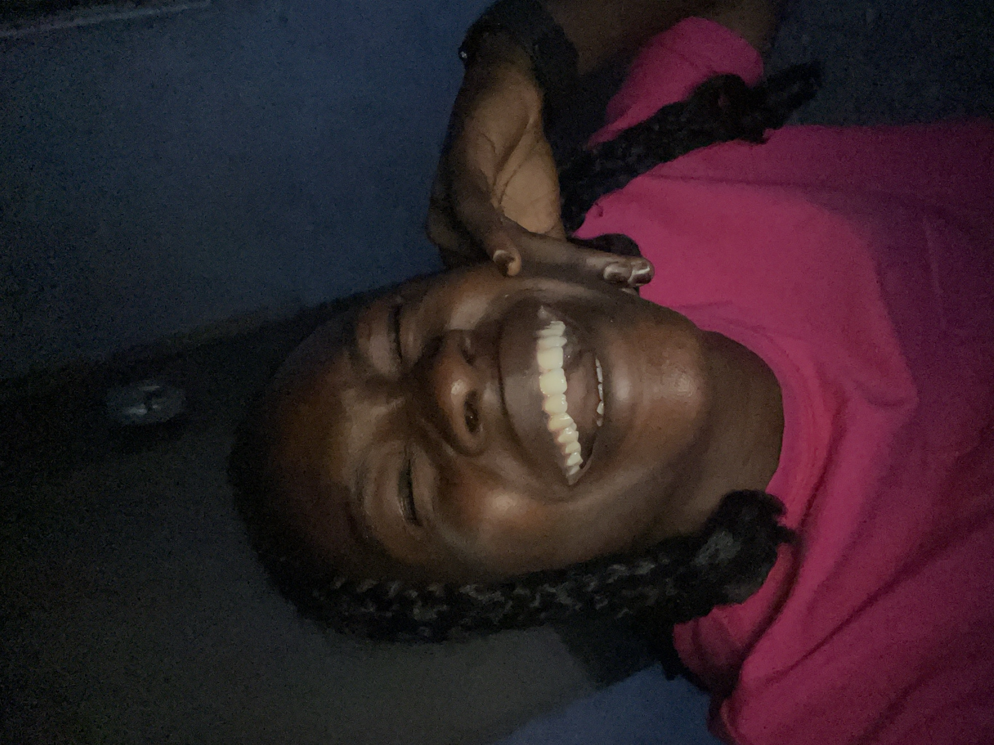 A close-up, head-and-shoulders shot shows a Black woman with braided hair, wearing a bright pink t-shirt, laughing with her eyes closed. Her right hand is raised to her face, touching her cheek. The background is dark and out of focus, with a hint of blue on the right side. The lighting is dim, with a spotlight effect on her face.