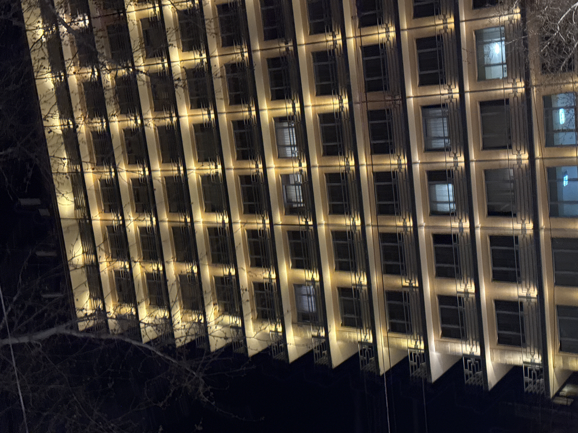Here is a description of the image:
This is a low-angle, nighttime shot of a multi-story building, likely in Yerevan, Armenia. The building has a grid-like facade with rows of windows. The building's exterior is illuminated with a warm, yellow light that accentuates the vertical and horizontal lines of its design. Some windows reflect interior lighting. There are bare tree branches in the foreground and near the top right of the frame. The background is dark, giving the building a stark contrast against the night sky. The image has a slightly tilted perspective.