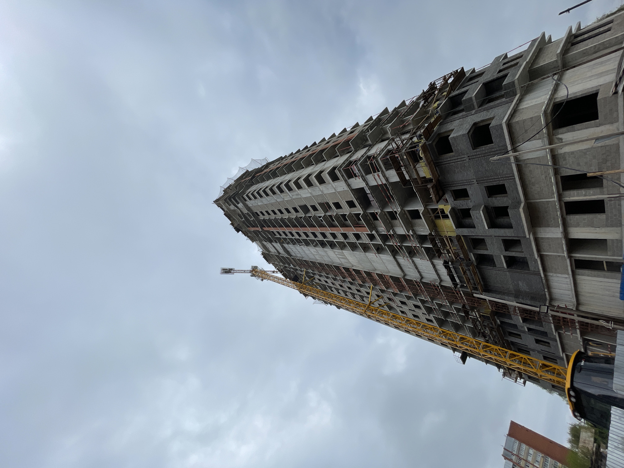 The image shows an unfinished high-rise building under construction against a cloudy sky. A yellow construction crane is attached to the side of the building, extending upwards. The building is located in Pervomaiskii, Russia. A bus is partially visible in the lower right corner, and another completed apartment building is visible in the lower center-right.