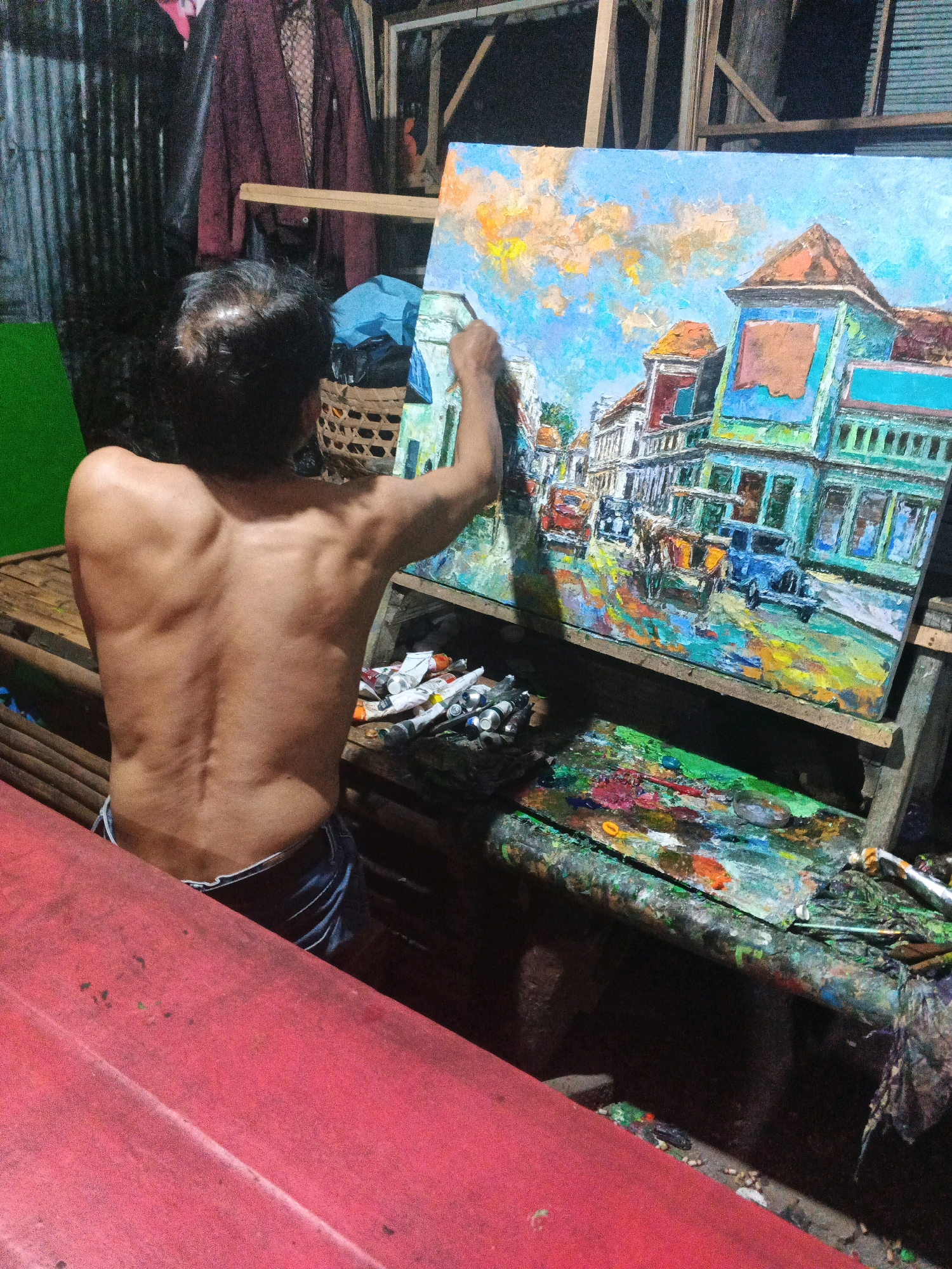An artist is painting a colorful cityscape on a canvas. The artist, a shirtless man with a muscular back, stands facing the canvas, holding a brush in his right hand and applying paint. The painting depicts buildings with orange rooftops and vibrant facades, along with vehicles and a street scene. The artist's work area is cluttered with tubes of paint, a palette covered in various colors, and a wicker basket. In the foreground, a red surface is visible, possibly a table or easel. The background is dark and appears to be an indoor space, with some fabric hanging and wooden structures. The overall setting suggests an artist's studio or workspace in a less formal environment. The painting itself is done in a thick impasto style, with visible brushstrokes adding texture and depth to the artwork. The lighting is dim, with artificial light illuminating the artist and his work. The image is a candid shot, capturing a moment of creative activity. The artist's back is the most prominent feature of the human subject. The cityscape in the painting is lively and detailed, with a sense of movement and activity. The presence of multiple paint tubes and the well-used palette indicate the artist's dedication to his craft. The overall impression is one of focused artistic endeavor in a somewhat rustic setting.