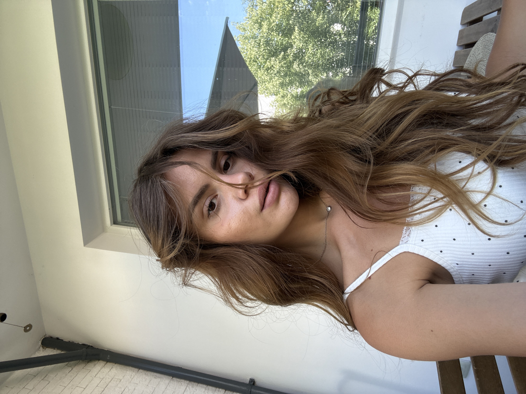 A young woman with long, wavy brown hair is pictured in a selfie from the chest up. She is wearing a white tank top with black polka dots and lace trim. A thin silver necklace with a small pendant rests on her collarbone. She is seated outdoors, with a white wall and a large window behind her. Through the window, green trees and a bright blue sky are visible. A drainage pipe is attached to the wall on the left side of the image. The background suggests an urban setting, potentially a balcony or rooftop patio. No specific Plovdiv, Bulgaria landmarks are identifiable.
