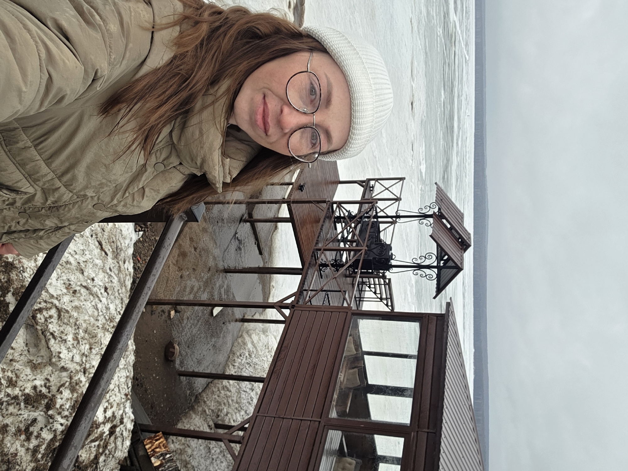 The image shows a selfie of a woman with red hair and glasses wearing a beige jacket and white beanie, taken at a gazebo on the edge of a body of water (likely a river or lake) in Chaikovsky, Russia. The woman is positioned to the left of the frame. The gazebo is constructed of wood and iron, with a small covered seating area. In the background, there is a partly frozen body of water, with a grey sky indicating it is likely winter. There is snow near the structure.