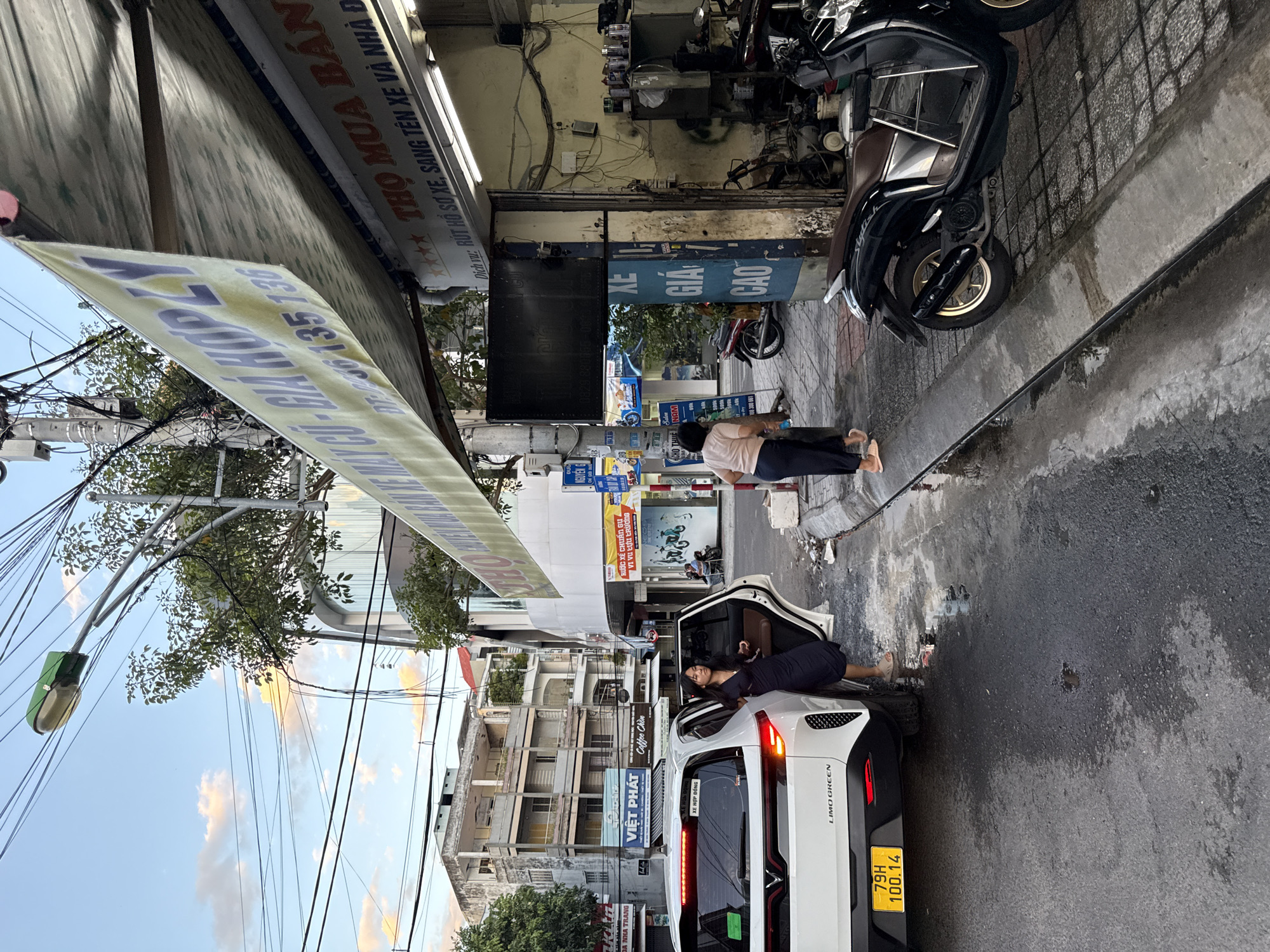 The image depicts a street scene in Nha Trang, Vietnam, likely in an urban commercial area. The time appears to be late afternoon or early evening, with the sky showing diffused light and some clouds.
In the foreground, a white electric vehicle, identified by the "LIMOGREEN" badging and a Vietnamese license plate reading "79H 100.14", is parked partially on the sidewalk. The driver's side door is open, and a young woman in a dark dress stands beside it, looking towards the right. Her posture suggests she might be waiting or observing something.
To the right of the car, a narrow sidewalk runs alongside a shopfront. A woman in a light-colored shirt and dark pants is seen walking away from the viewer, towards the shop. She is carrying a light-colored bag.
The shop itself appears to be a repair or service establishment, indicated by the presence of tools and equipment visible within its open doorway, as well as a large banner above it. The banner, predominantly yellow, displays Vietnamese text and phone numbers, advertising services related to vehicles. A sign further right, also in Vietnamese, mentions "THO MUA BÁN XE, SANG TÊN XE VÀ NHÀ ĐẤT," suggesting services related to buying/selling vehicles and property.
Several motorbikes are parked along the sidewalk in front of the shop, including a black scooter in the immediate foreground. The street is wet, indicating recent rain or cleaning. Overhead, a complex network of electrical wires crisscrosses the sky, a common feature in Vietnamese urban landscapes. In the background, multi-story buildings line the street, characteristic of a city. One building has a curved glass facade. Another building displays signage for "VIET PHAT" and "COFFEE CHIN." A smaller blue sign indicates "DU LICH HUE," suggesting tourism-related services might be nearby. The overall impression is one of a busy, functional street in a Vietnamese city.