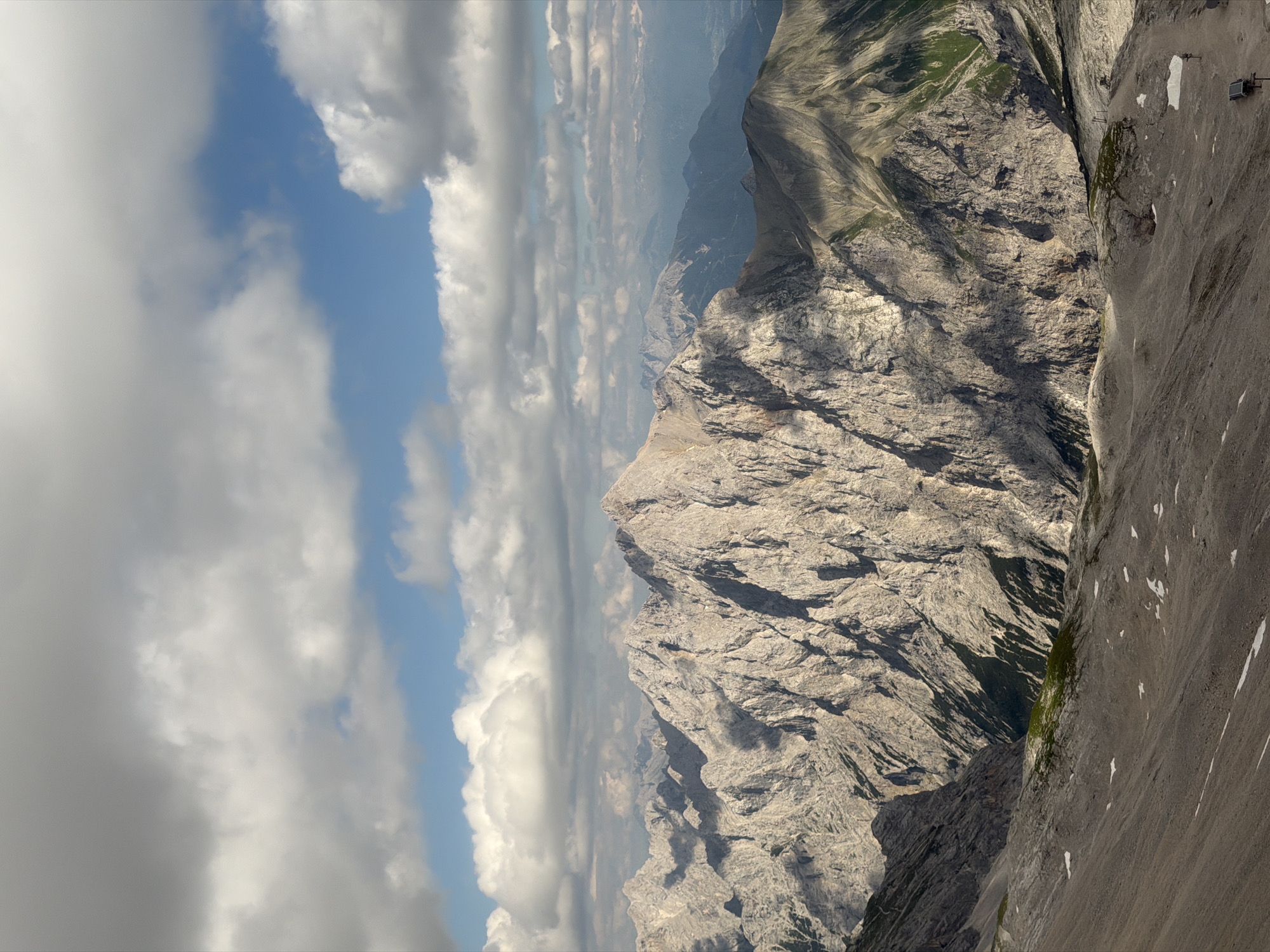 The image shows a panoramic view of a mountainous landscape, likely taken from a high vantage point. The scene is dominated by rugged, rocky mountain peaks and slopes. In the foreground and mid-ground, the mountains are characterized by their steep, textured surfaces, with visible rock formations and scree. Patches of green vegetation, appearing as grassy slopes or moss, are interspersed with the rock, especially in shadowed areas or on less steep inclines.
The sky above is partly cloudy, with a mix of bright blue patches and large, fluffy white and gray clouds. The lighting suggests it is daytime, likely late morning or early afternoon, given the angle of the sunlight creating distinct shadows on the mountains. The weather appears to be fair, though the presence of clouds indicates variable conditions.
In the far distance, additional mountain ranges are visible, fading into a hazy blue, suggesting a significant depth to the landscape. The overall impression is one of grandeur and natural beauty.
On the right side of the image, on one of the steeper rocky surfaces, there are some man-made objects. These appear to be fixed points or anchors, possibly part of a climbing route or a safety installation. Small white patches on the rock face might be snow or ice, though their size and distribution suggest they are unlikely to be significant snowfields.
There is no visible text in the image. The setting is clearly a high alpine environment, consistent with mountainous regions like Garmisch-Partenkirchen, Germany. The perspective suggests the viewer is either at a very high elevation on a mountain or possibly in an aircraft or on a high cable car. There are no people or animals explicitly visible, emphasizing the wild and remote nature of the location.