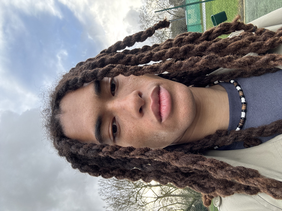 The image is oriented sideways, with the subject's head positioned on the left side of the frame. It features a close-up portrait of a person, likely male, with long, thick, dark brown dreadlocks that frame their face and extend across the image.
The person has a medium-dark skin tone and is looking directly at the camera with dark, open eyes. Their eyebrows are dark and well-defined. They have a straight nose and full, naturally pink-hued lips that are slightly parted. Subtle stubble is visible on their chin and upper lip area.
Around their neck, they wear a necklace composed of small, cylindrical beads in alternating black, white, and brown (or wooden) colors. They are dressed in a dark blue or grey crew-neck top, partially covered by a light-colored, possibly beige or light olive green, jacket or coat, a button of which is visible on the far right side of the frame.
The background indicates an outdoor setting on an overcast day. The upper part of the image shows a cloudy sky with some brighter patches. Bare branches of deciduous trees are visible in the mid-ground, suggesting it might be late autumn or winter. In the upper right corner of the frame (as oriented), a green mesh fence, possibly from a tennis court or sports field, can be seen, along with a dark green object, perhaps a bench or trash can, behind it.
The lighting is soft and diffused, characteristic of an overcast day. The person's expression is neutral and calm. There is no visible text in the image.