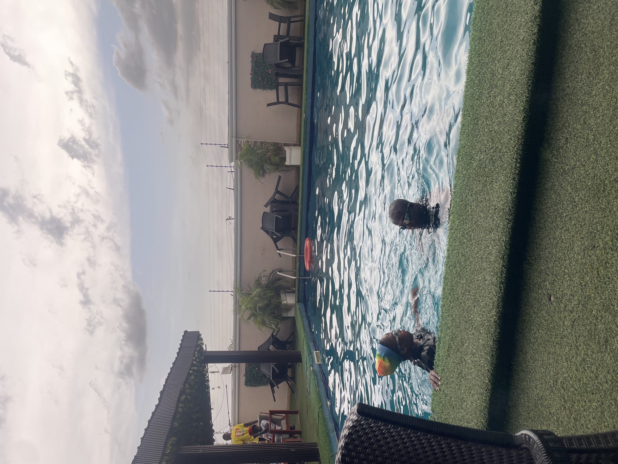 The image displays an outdoor swimming pool area, likely at a hotel or private residence in Abuja, Nigeria, under a partly cloudy daytime sky. The scene is oriented vertically in the provided image but depicts a horizontal landscape.
In the main section of the frame, a swimming pool with clear, rippling blue water is visible. Two individuals are in the water: one person, closer to the foreground, appears to be swimming on their back and is wearing a colorful, possibly rainbow-striped, swim cap. Another person is further into the pool, with only their head visible, wearing a darker swim cap or having dark wet hair. A red lifebuoy floats in the pool.
The area immediately bordering the pool is covered with artificial green turf. Beyond the turf and pool, against a light-colored wall, several dark outdoor chairs and tables are arranged. Potted plants are strategically placed along the wall, adding decorative elements.
On the left side of the frame, a dark, slatted pergola or awning structure provides shade. Beneath this structure, a person wearing a bright yellow t-shirt is seated at a table. The text or logo on their yellow shirt is not discernible. In the immediate foreground, part of a dark, woven outdoor furniture item, possibly a chair or table, is visible.
The overall atmosphere suggests a leisurely and relaxed setting, with people enjoying the pool on a bright but partly overcast day.