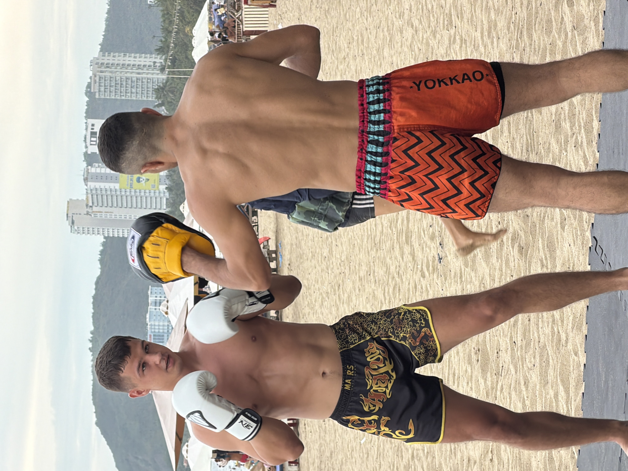 Two men are engaged in Muay Thai training on a sandy beach in Nha Trang, Vietnam. The man in the foreground, facing the viewer, wears white boxing gloves and black Muay Thai shorts with gold and red tribal patterns. His training partner, with his back to the camera, wears yellow focus mitts and orange Muay Thai shorts with a black zigzag pattern on the left leg and "YOKKAO" written vertically on the right. In the background, several white buildings are visible, suggesting an urban coastal setting. The sky is overcast.