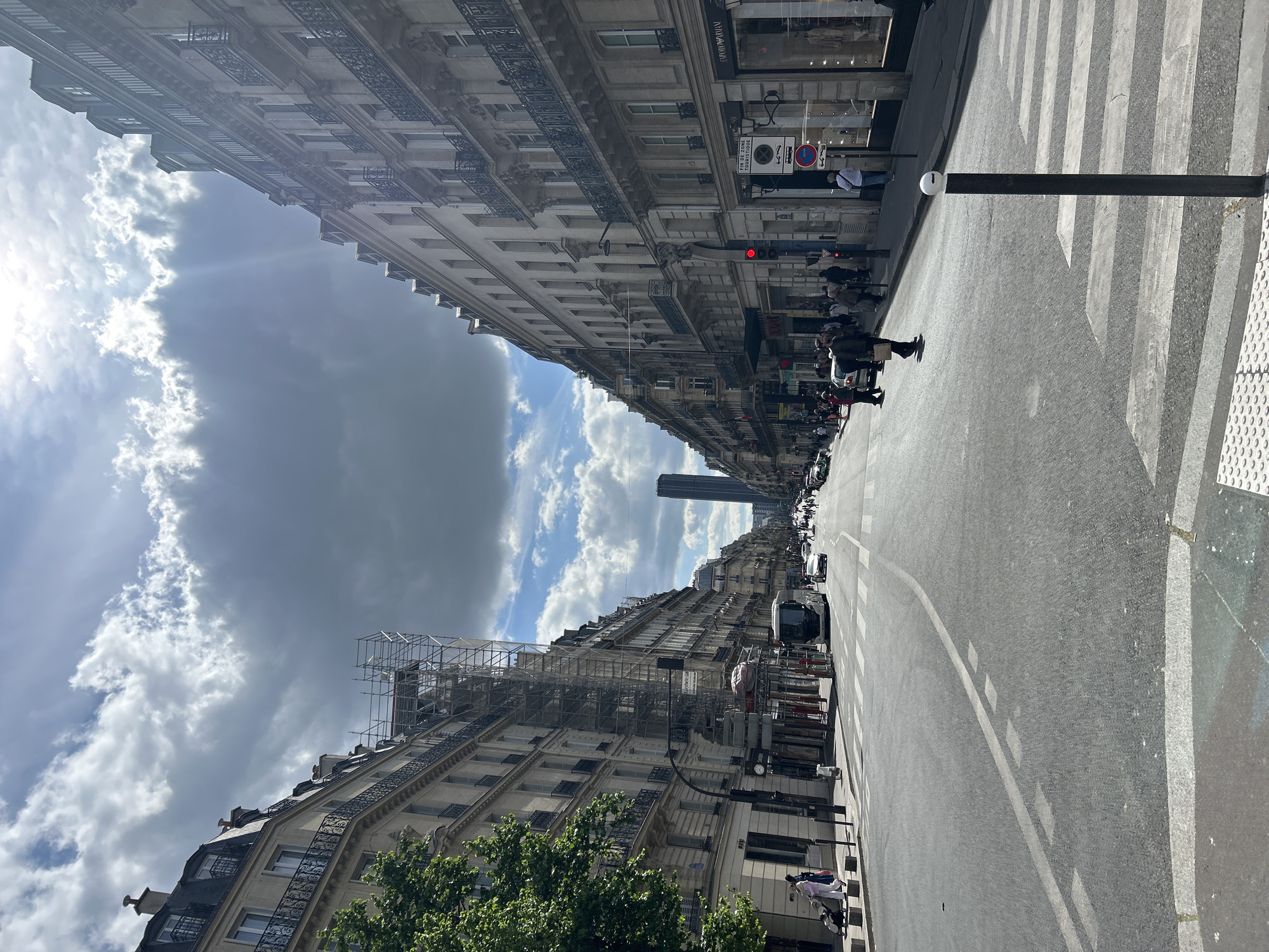 A street view shows a city street lined with tall buildings on either side, and a road runs through the center. The perspective suggests the image was taken at street level. The sky above is filled with a mix of light and dark clouds. At the far end of the road, a tall skyscraper is visible. People are present on the sidewalks, and cars are parked along the street. The overall impression is an urban setting, possibly a European city, on a partly cloudy day.