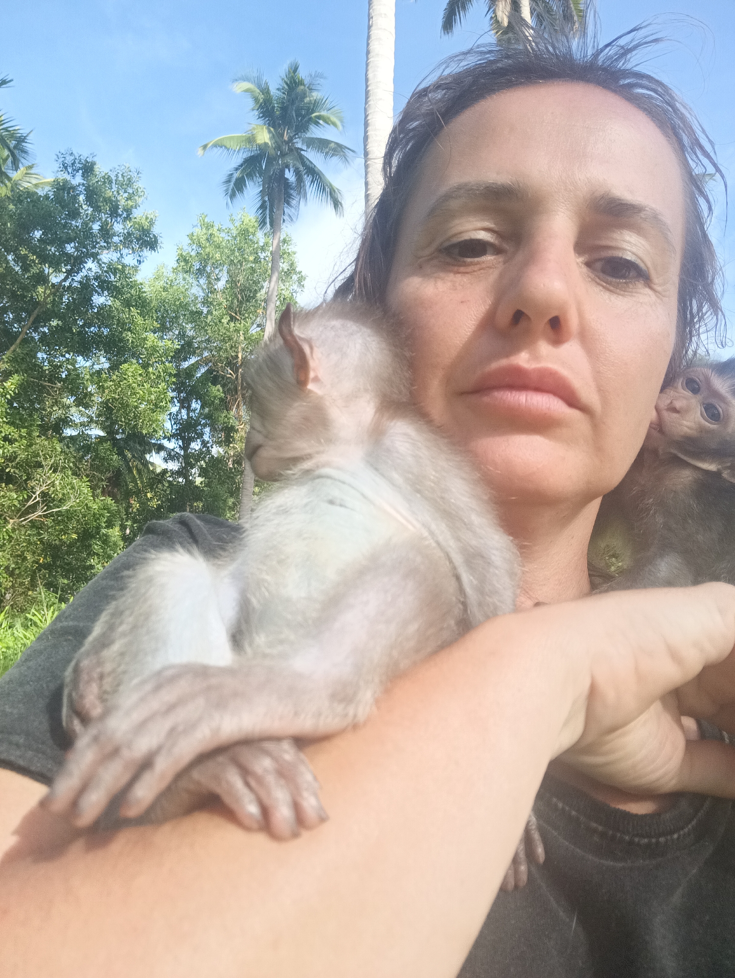 A woman is holding two baby monkeys in what appears to be a tropical outdoor setting, likely in the city of Baan Thong Nang, Thailand. She has one monkey perched on her shoulder and held with her arm, and another peeking out from behind. The background includes trees and a light blue sky.