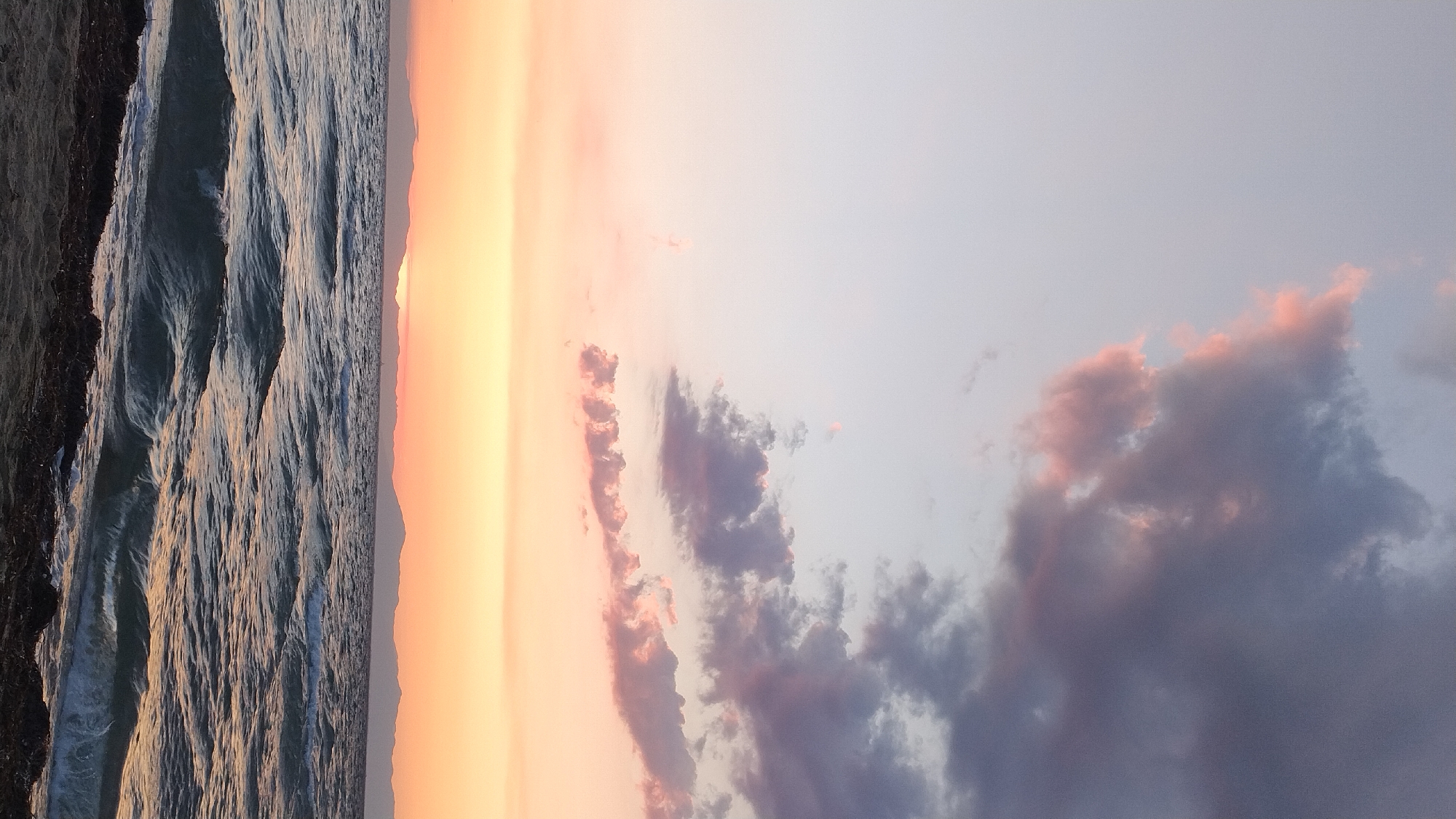 The image depicts a sunset over the Aegean Sea, viewed from the beach in Neoi Epivates, Greece. The sky transitions from a pale blue at the top to hues of pink, orange, and yellow near the horizon. Clouds, tinged with pink and dark gray, are scattered across the sky. The sea is choppy, with visible waves and whitecaps. In the distance, a silhouetted mountain range stretches across the horizon, with the sun partially obscured behind it, casting a soft glow. The foreground shows a sandy beach with some seaweed. No people or animals are visible in the scene.