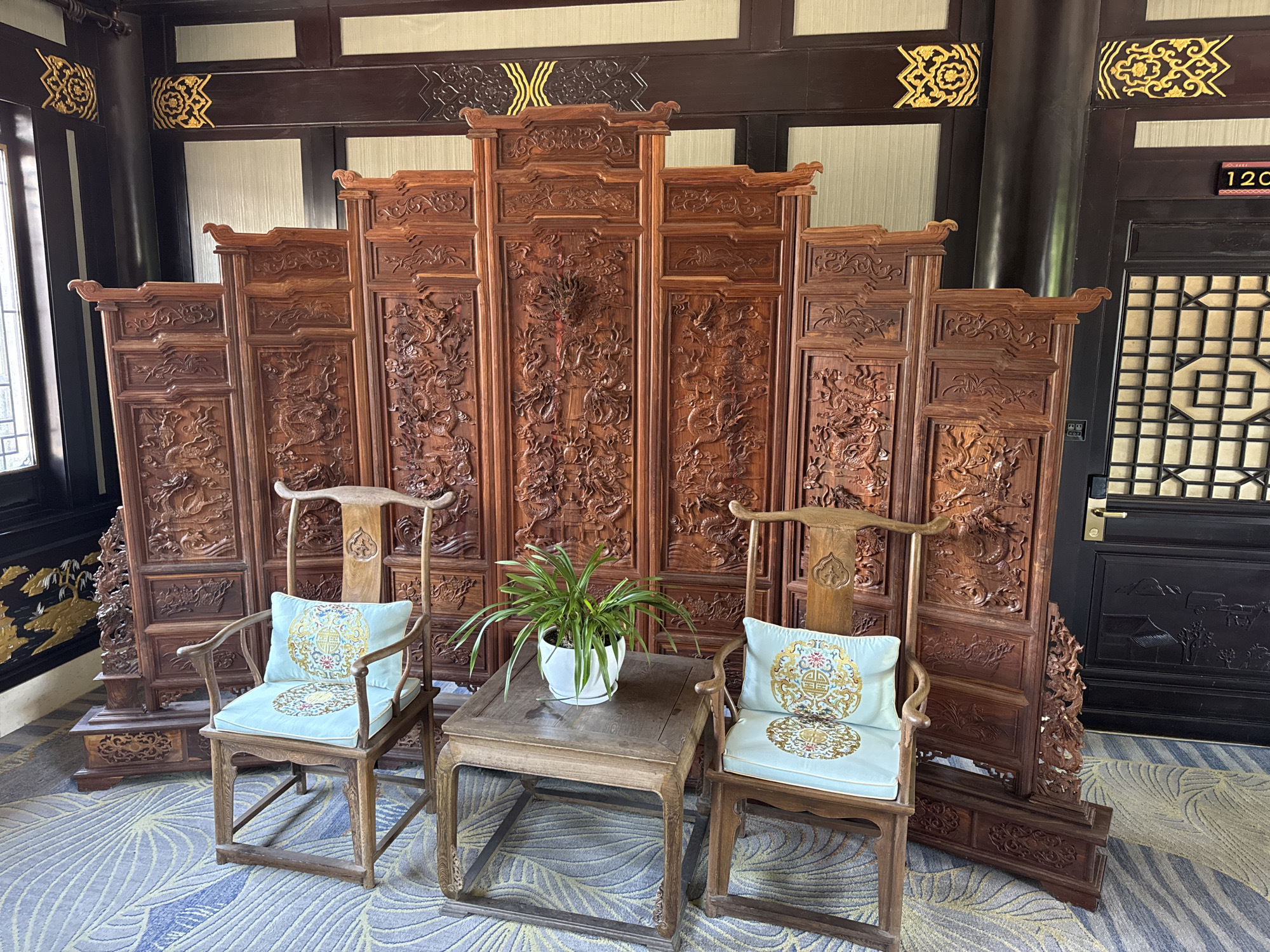 In a room with dark wood paneling and a patterned carpet, two ornate wooden chairs flank a small wooden table. The chairs are light-colored wood with curved arms and backrests, each adorned with a light blue cushion embroidered with gold circular designs. The table is also made of wood and holds a small potted plant with green leaves. Behind the furniture, a large, intricately carved wooden screen stands, featuring detailed dragon motifs. The room appears to be in a traditional Chinese architectural setting. The image is a still photograph of this indoor scene.