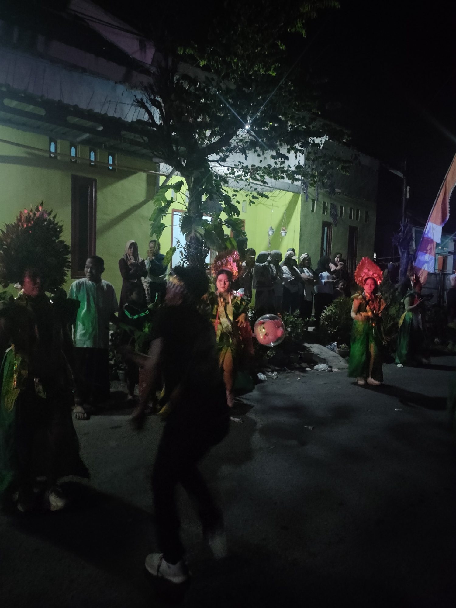 A group of people are gathered on a street at night in Tongaswetan, Indonesia, for what appears to be a performance or festival. Several individuals are dressed in elaborate, traditional costumes, some with tall, feathered headdresses and brightly colored, flowing garments. One person in the foreground is in motion, their body blurred by movement, suggesting they are dancing. Other figures in the background are observing the activity. The setting includes a yellow building with windows and a roof, a tree with dense foliage, and a flag on a pole. The ground is paved, and there are some discarded items and debris scattered around. The lighting is dim, with some artificial lights illuminating parts of the scene, creating shadows.