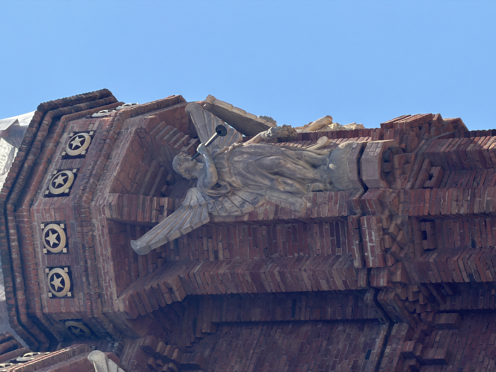 The image shows a close-up, low-angle view of a detailed architectural facade against a clear blue sky, suggesting daytime. The facade is constructed primarily of reddish-brown bricks, arranged in intricate patterns and layered to create a textured, almost sculptural effect.
Dominating the center of the frame is a relief sculpture of a winged figure, likely an angel, lying down and playing a horn. The figure is carved from a lighter material, possibly stone or concrete, and appears aged and weathered. The angel's body is draped in flowing robes, and one wing is prominently visible. The horn is depicted as a simple, narrow tube. The sculpture is integrated into the brickwork, appearing as if emerging from it.
To the left of the sculpture, the brickwork features a column of square decorative elements, each containing a stylized five-pointed star within a circular border, set against a golden background. These star motifs are repeated at various levels of the facade. The overall architectural style appears historic, with a strong emphasis on ornamentation and craftsmanship. The setting is likely an exterior building in Barcelona, Spain, given the context, possibly a church, monument, or an older public building known for its distinctive architectural features. The weather appears clear and sunny, typical of a pleasant day in Barcelona. There is no visible text in the image.
