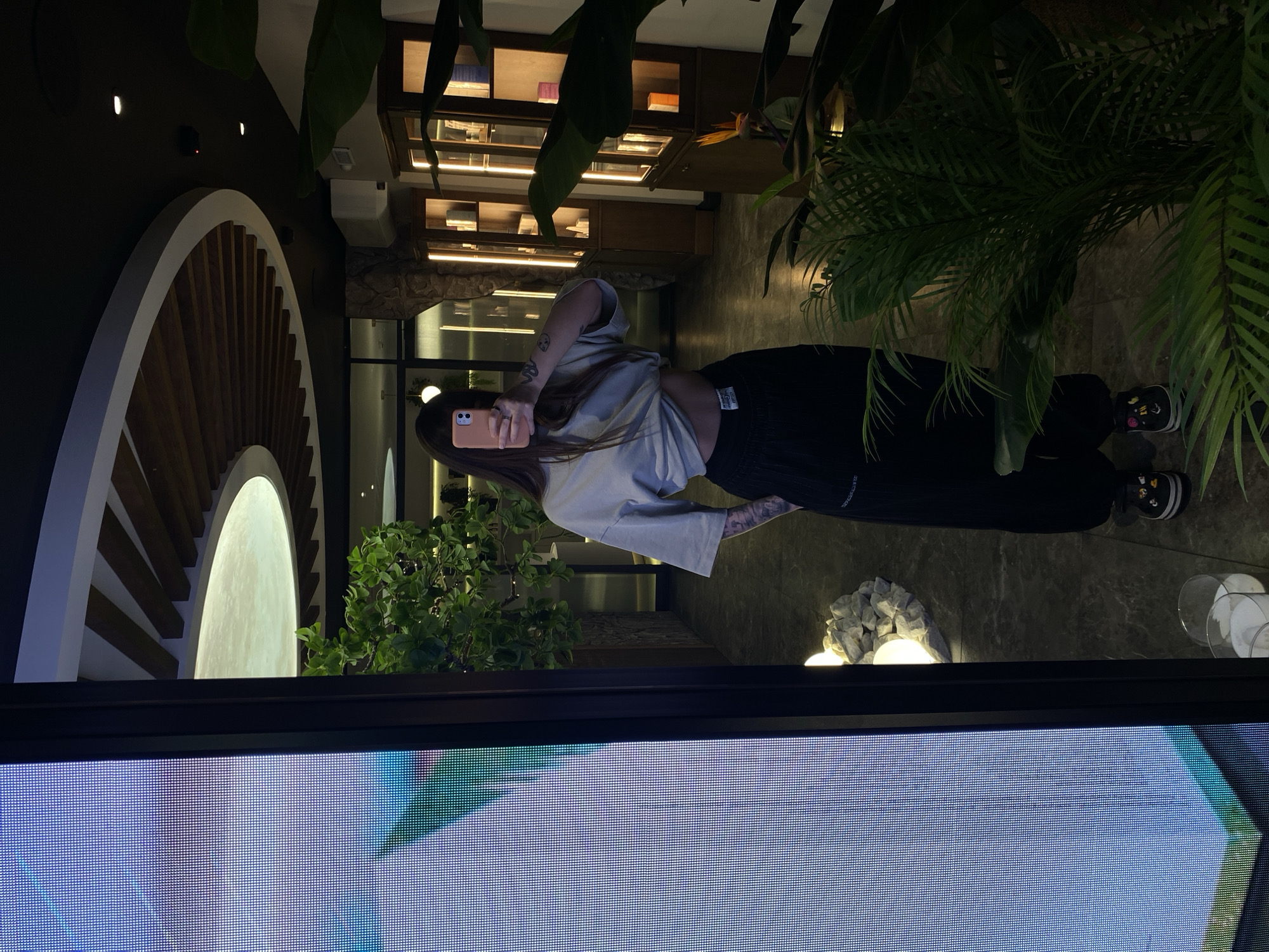 A woman with long brown hair, wearing a beige t-shirt and black pants, stands in a brightly lit interior space, holding a pink smartphone up to take a photo. Tattoos are visible on her arm. Behind her is a decorative wall element with a circular, arched design. Lush green plants are placed throughout the space, contributing to a modern, somewhat tropical ambiance. In the background are shelves with various objects and indirect lighting. The scene is partially obstructed by what appears to be the lower edge of a large screen displaying an indiscernible image. The location details in Votkinsk, Russia, are not evident from the provided image.