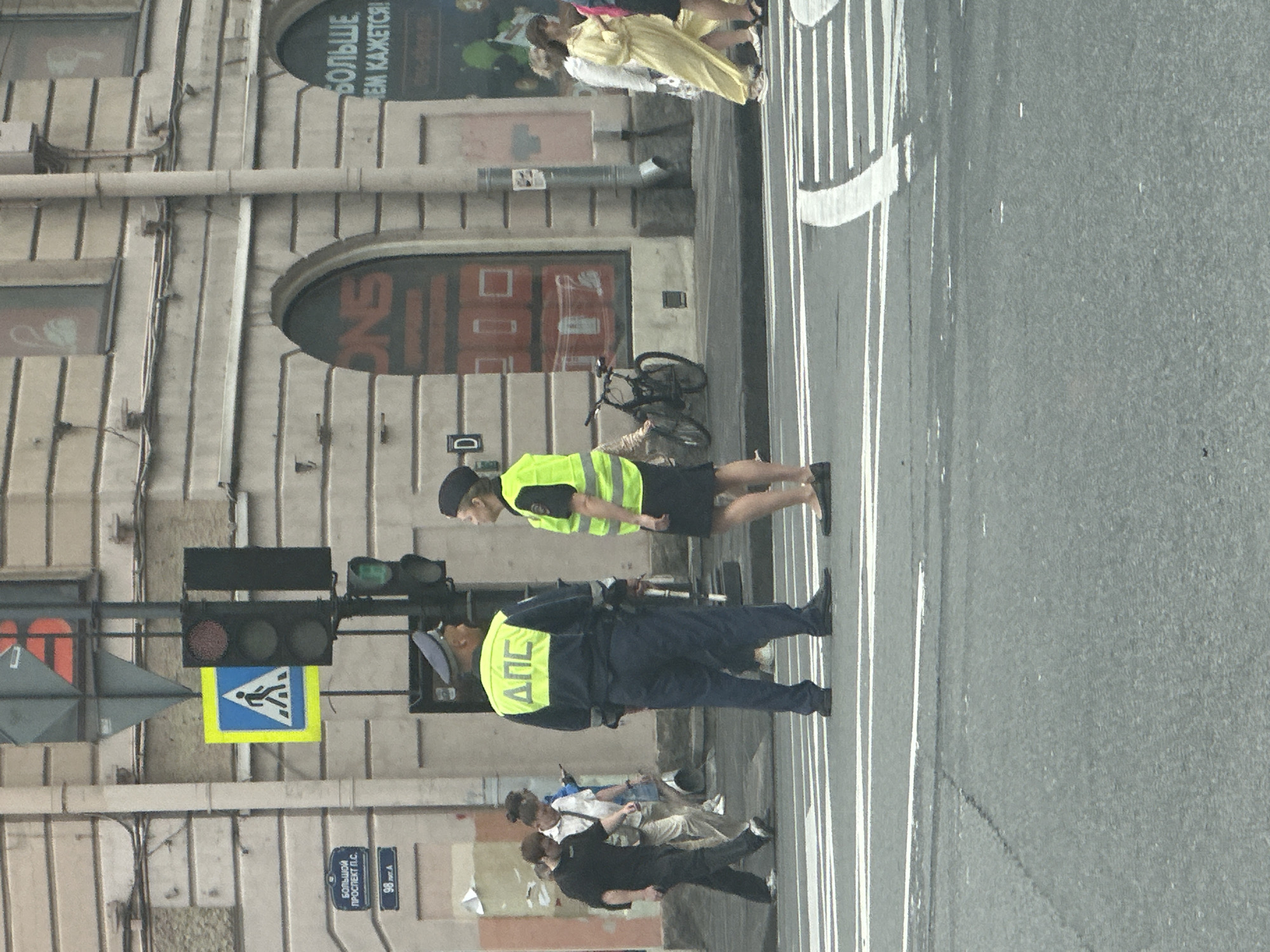 The image captures a street scene in Saint Petersburg, Russia, likely near a pedestrian crossing, given the presence of traffic lights and pedestrian signage. Two individuals in official-looking uniforms, one male and one female, are standing at the intersection, appearing to be engaged in a conversation. Both are wearing reflective vests over their uniforms. The male officer wears a dark blue uniform with a cap and a yellow vest with the Cyrillic letters "ДПС" prominently displayed. The female officer is also in uniform with a black skirt and a yellow vest.
In the background, several other pedestrians are visible, some walking across the street and others on the sidewalk. A bicycle is parked near the building on the right. The architecture suggests an urban environment with a light-colored, multi-story building forming the backdrop. Signage on the building and lampposts, including a street sign indicating "БОЛЬШОЙ ПРОСПЕКТ П.С." and a number "98", help to identify the location. Traffic lights, some with red lights illuminated, are also present, indicating active traffic management. The street itself is asphalt with white pedestrian crossing markings. The overall lighting suggests it is daytime, possibly overcast.