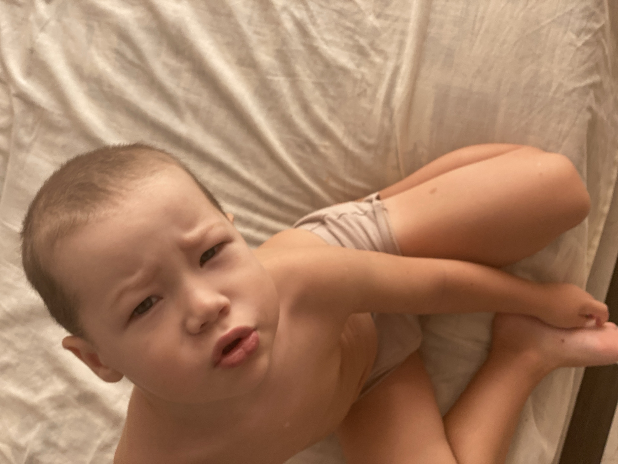 This is a close-up, slightly angled, indoor photograph of a young child lying on a light-colored, textured surface, possibly a bed or sofa. The child is facing towards the camera with a pensive or questioning expression, their mouth slightly open. Their hair is short and light brown, and they have light skin with some freckles visible on their arms and legs. They are wearing light-colored underwear. The child's body is positioned with their legs bent and their arms crossed in front of them. The lighting appears soft, suggesting an indoor environment, possibly during the day. There is no visible text in the image. The context provided indicates the location as Sochi, Russia, but there are no specific visual cues within the image to confirm this.