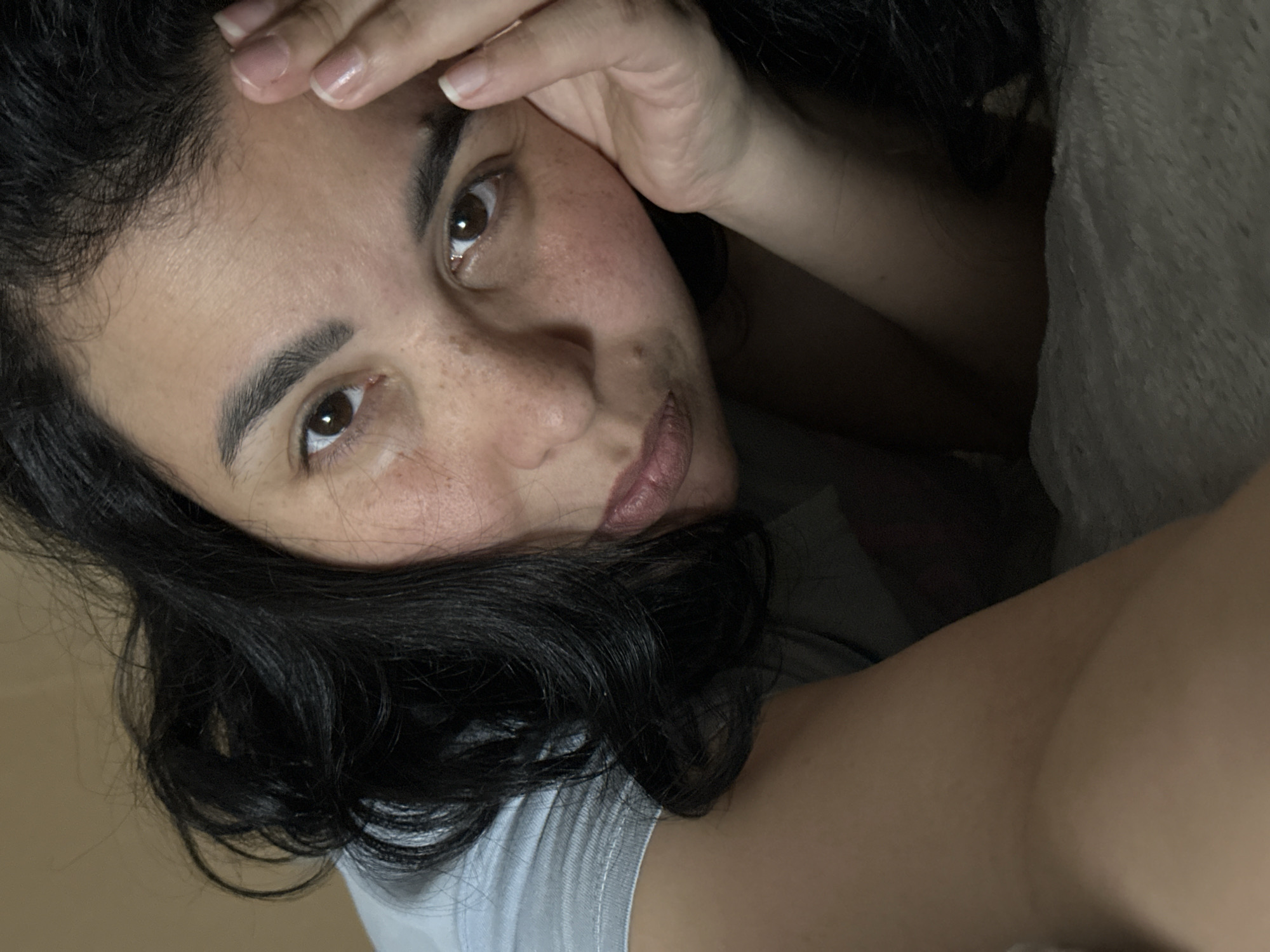 A close-up, angled selfie of a woman with dark, wavy hair. Her skin has visible freckles. She has brown eyes and her lips are slightly parted. Her hand is resting on her forehead. She is wearing a light-colored shirt. Part of a gray blanket or pillow is visible in the background. The location is unspecified.