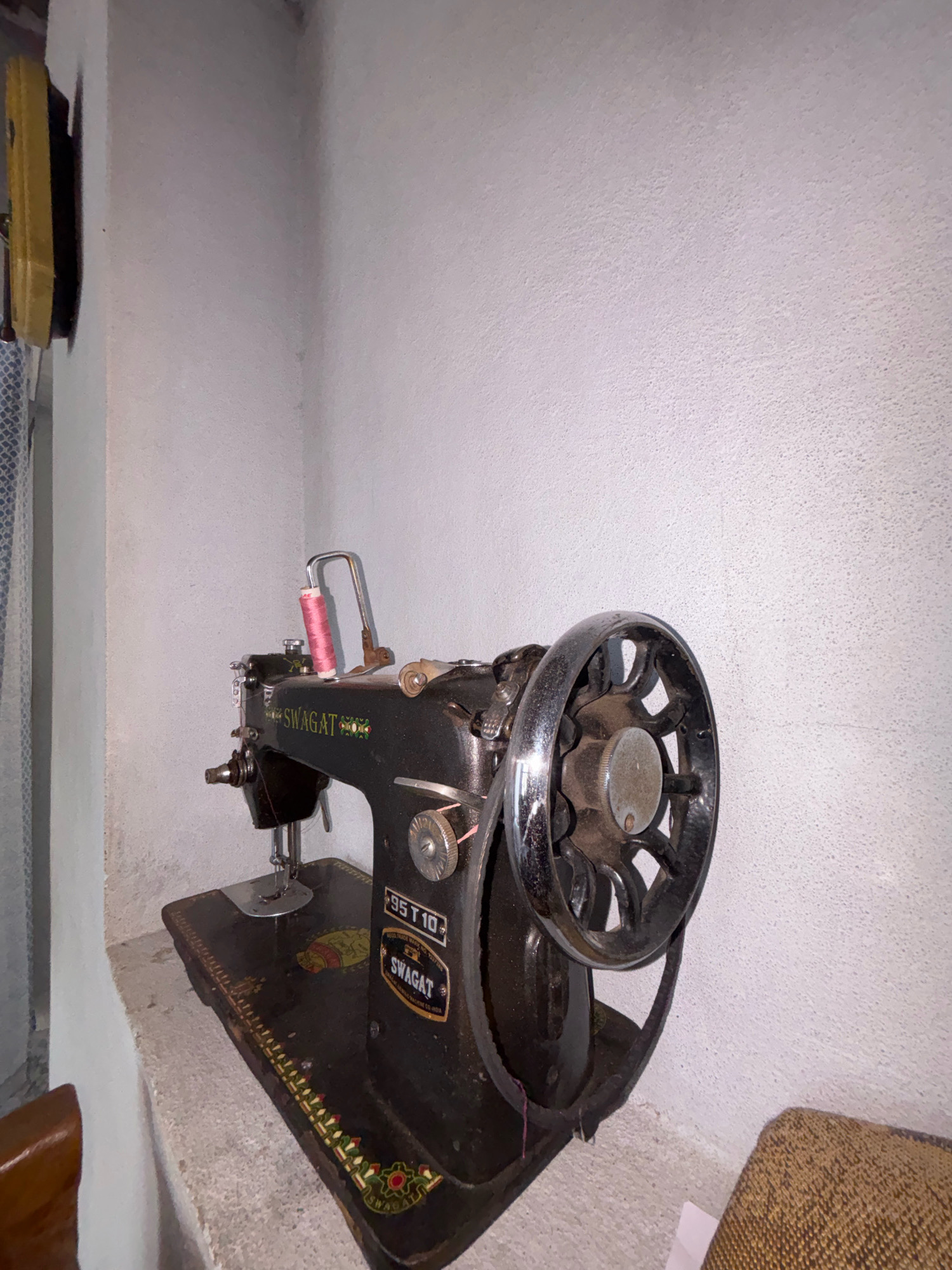 An old black manual sewing machine, identified by the "SWAGAT" logo, is placed on a rough, gray concrete ledge. A spool of pink thread is positioned on a metal spool pin at the top of the machine. To the left of the sewing machine, a glimpse of a patterned blue and white fabric and a yellow object are visible. The background consists of a rough, white textured wall. This scene is located in Ghataprabha, India.