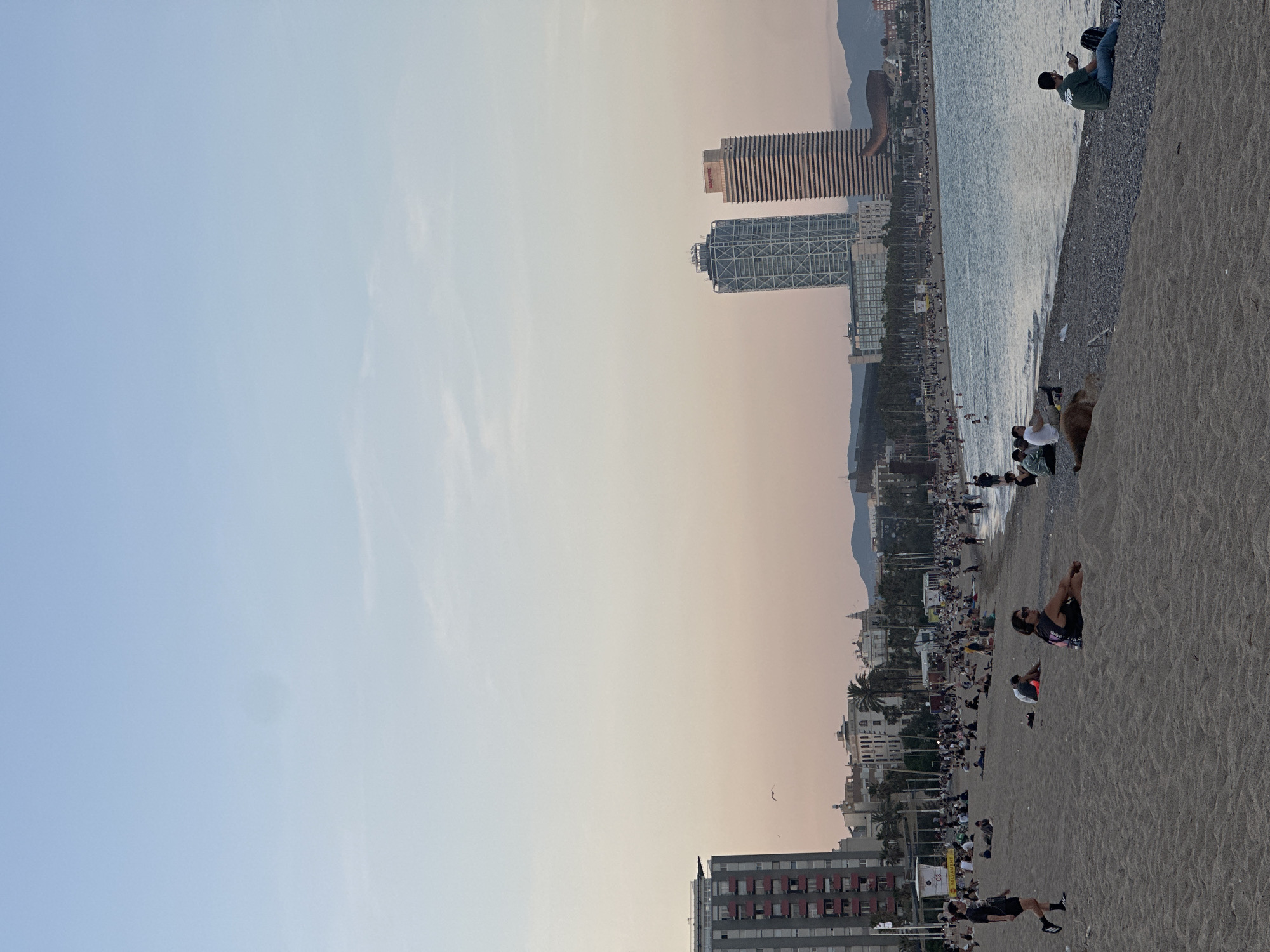 The image captures a scene at Barceloneta Beach in Barcelona, Spain. The beach is densely populated with people engaged in various activities, such as sunbathing, walking, and sitting by the sea. In the background, the skyline features prominent buildings, including the Hotel Arts and Torre Mapfre. The coastline is visible, and mountains appear in the far distance. The sky transitions from blue to a soft sunset hue. The foreground is dominated by the sandy beach, showing a range of textures and depths.