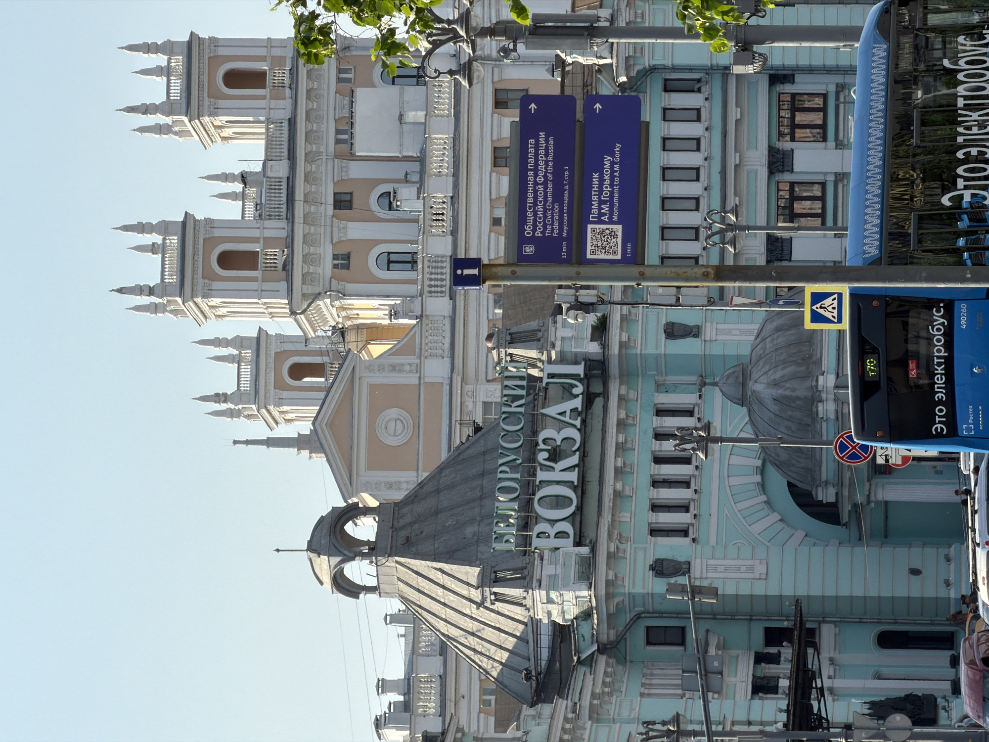 Here is a description of the image:
The image shows a view of the Belorussky railway station in Moscow, Russia. The station building is prominent in the image, featuring its architectural details, including towers and the sign with "Белорусский Вокзал" (Belorussky Station) in Russian. There are signs giving directions to the Civic Chamber of the Russian Federation and a monument to A.M. Gorky. A blue electric bus is seen on the right side of the image. A pedestrian crossing sign is also visible. The photo appears to be taken from a street level perspective on a clear day.