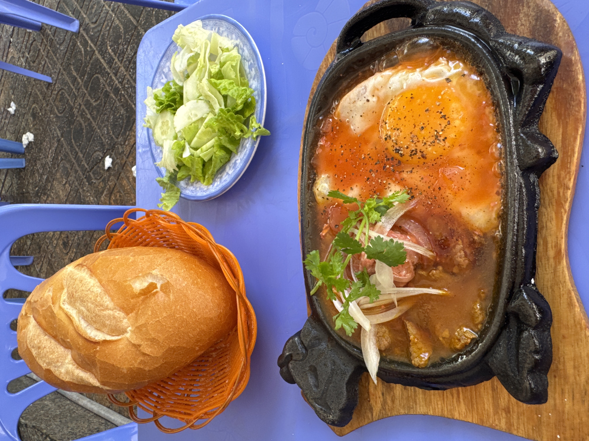 An overhead shot shows a meal on a blue table, likely at a street-side restaurant in Bac Nha Trang Ward, Vietnam. In the foreground, a sizzling black cast-iron skillet, shaped like a bull, holds a dish of eggs, meat, and vegetables in a savory sauce, topped with cilantro and onion. A side of fresh lettuce and cucumber is on a small plate to the right. To the left, a crusty baguette sits in an orange basket. The background reveals blue plastic chairs and a tiled walkway, suggesting an outdoor dining setting.