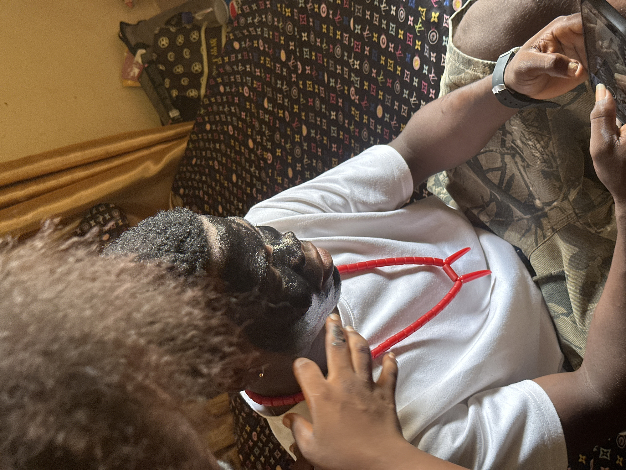 The image portrays an intimate indoor scene in Ikot Ekpene, Nigeria, featuring two individuals. The central subject is an adult male, viewed from an overhead perspective, reclining on a surface covered with a dark, richly patterned fabric resembling a Louis Vuitton monogram. He has dark skin, black hair, and wears a white short-sleeved shirt. A prominent red beaded necklace, likely made of coral, rests around his neck and on his chest. He also wears camouflage shorts and a black smartwatch on his left wrist. With both hands, he holds a smartphone or tablet, appearing to be engaged with the device.
In the foreground, part of another person is visible, characterized by curly brown hair and a hand gently touching the adult male's face, suggesting a moment of affection or playful interaction. The adult male's gaze is directed upwards, potentially at the person touching him or towards the ceiling.
The setting appears to be a domestic space, possibly a bedroom or living room, indicated by the patterned fabric and a gold-colored curtain or fabric visible in the background. The lighting suggests daytime. The overall mood is relaxed and personal. No discernible text is visible within the image.