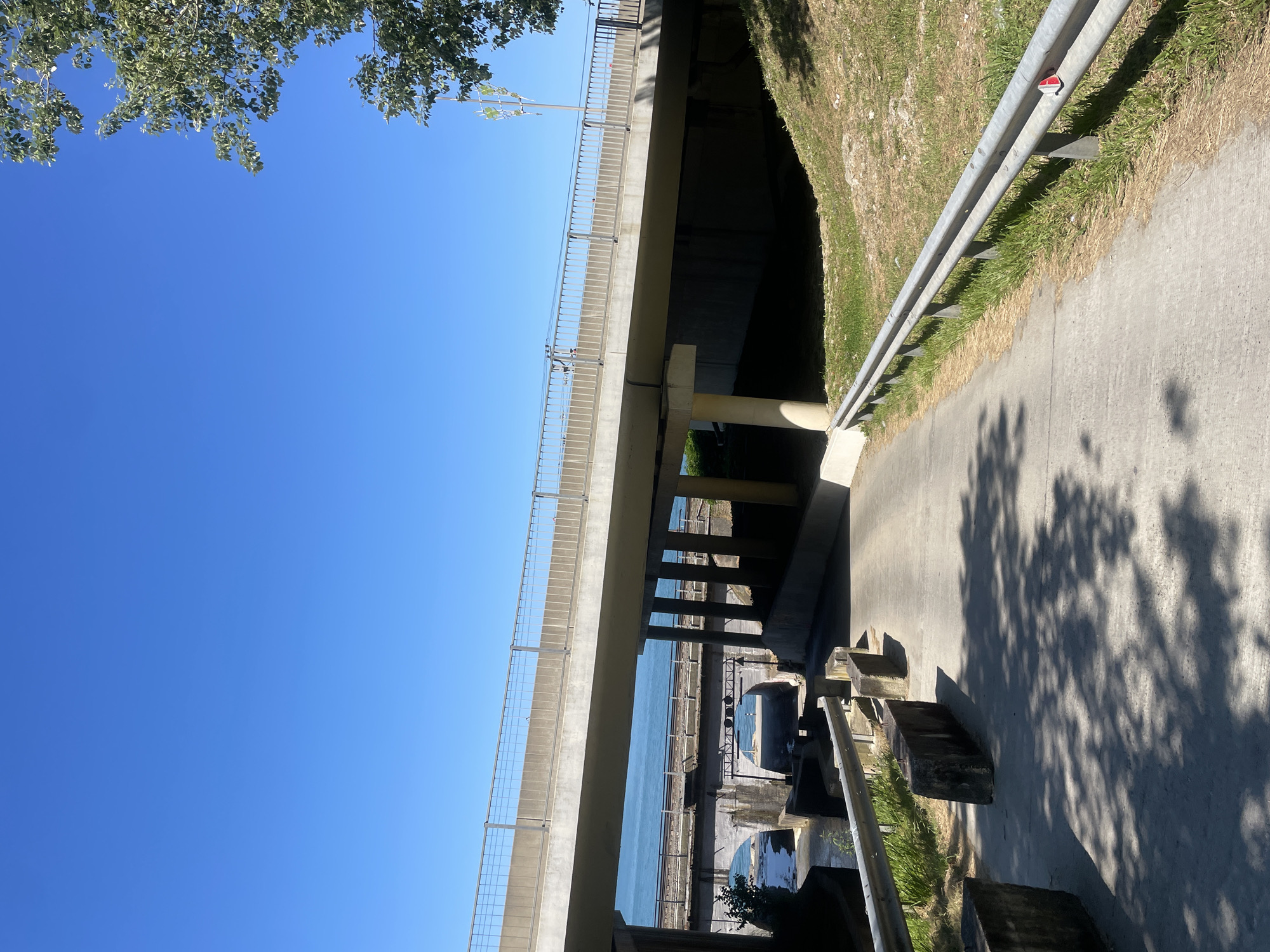 The image depicts a concrete bridge with a metal railing, viewed from a lower vantage point. The bridge spans over a body of water, likely a sea or large lake, with a waterfront area visible beneath it. There are some concrete structures and what appear to be railway tracks under the bridge. The sky is clear and blue, suggesting a sunny day. The lighting indicates it is daytime, possibly late morning or afternoon, given the clear shadows cast on the concrete path. The scene appears to be outdoors, possibly a recreational or public space near a waterfront. There is some vegetation, including trees, visible in the upper left corner. A small red and white sticker is visible on the metal railing. No people are clearly visible. The overall atmosphere is calm and bright.