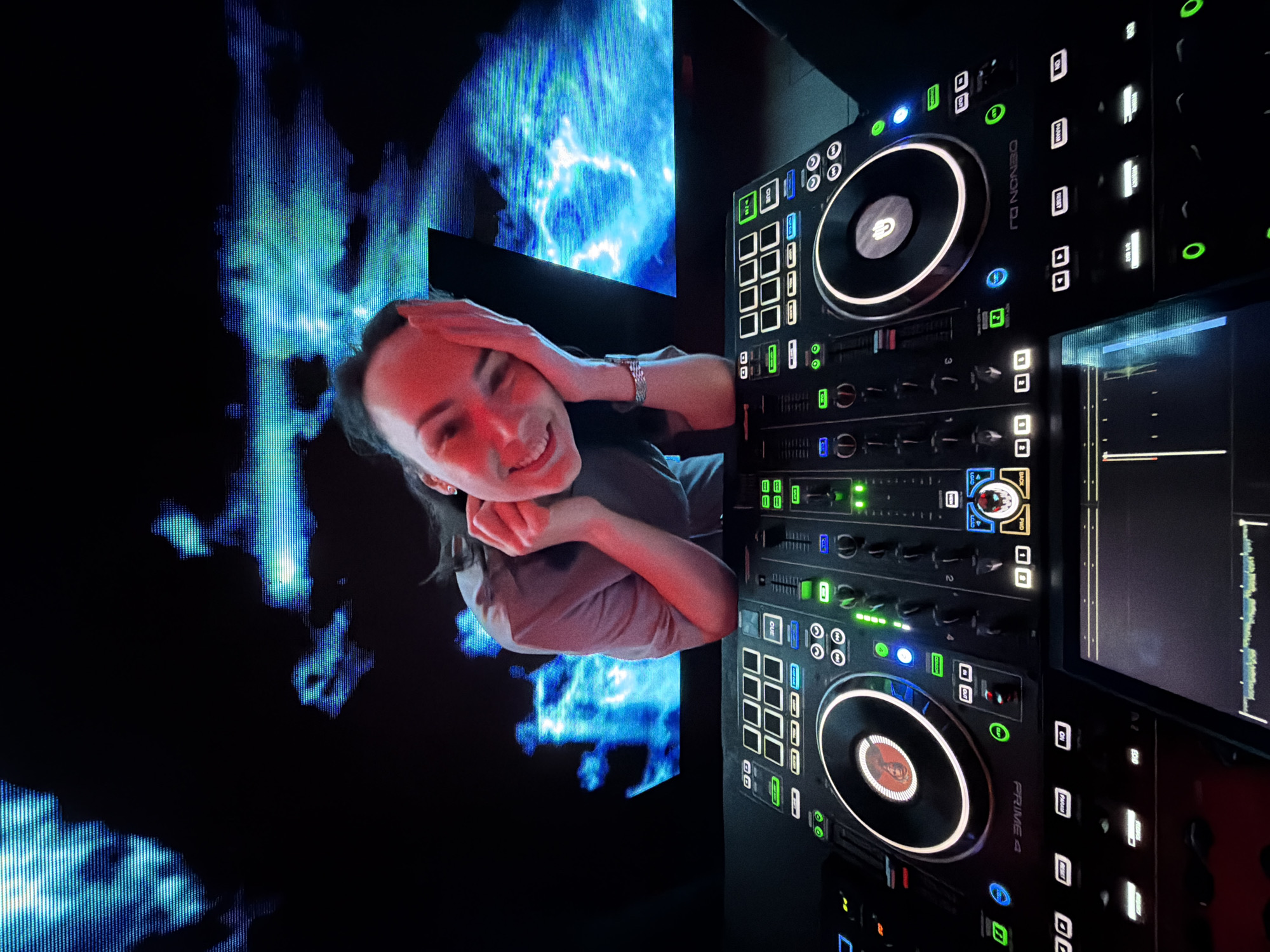 Here is a description of the image:
The image shows a woman with dark hair smiling and posing in front of a DJ setup and a vibrant, illuminated backdrop. The woman is wearing a light-colored shirt and has her hands near her face in a relaxed pose. To her right, the DJ equipment is prominently displayed, featuring various knobs, buttons, and illuminated elements. The equipment appears to be a Denon DJ Prime 4. Behind the woman is a large display screen exhibiting dynamic patterns and colors, mainly blues and greens, which create a lively atmosphere. The overall lighting is low, highlighting the illuminated parts of the DJ equipment and the display screen. The image seems to be taken in an entertainment setting or a club environment. Votkinsk, Russia is not directly visible in the image, nor can it be inferred from the image contents.