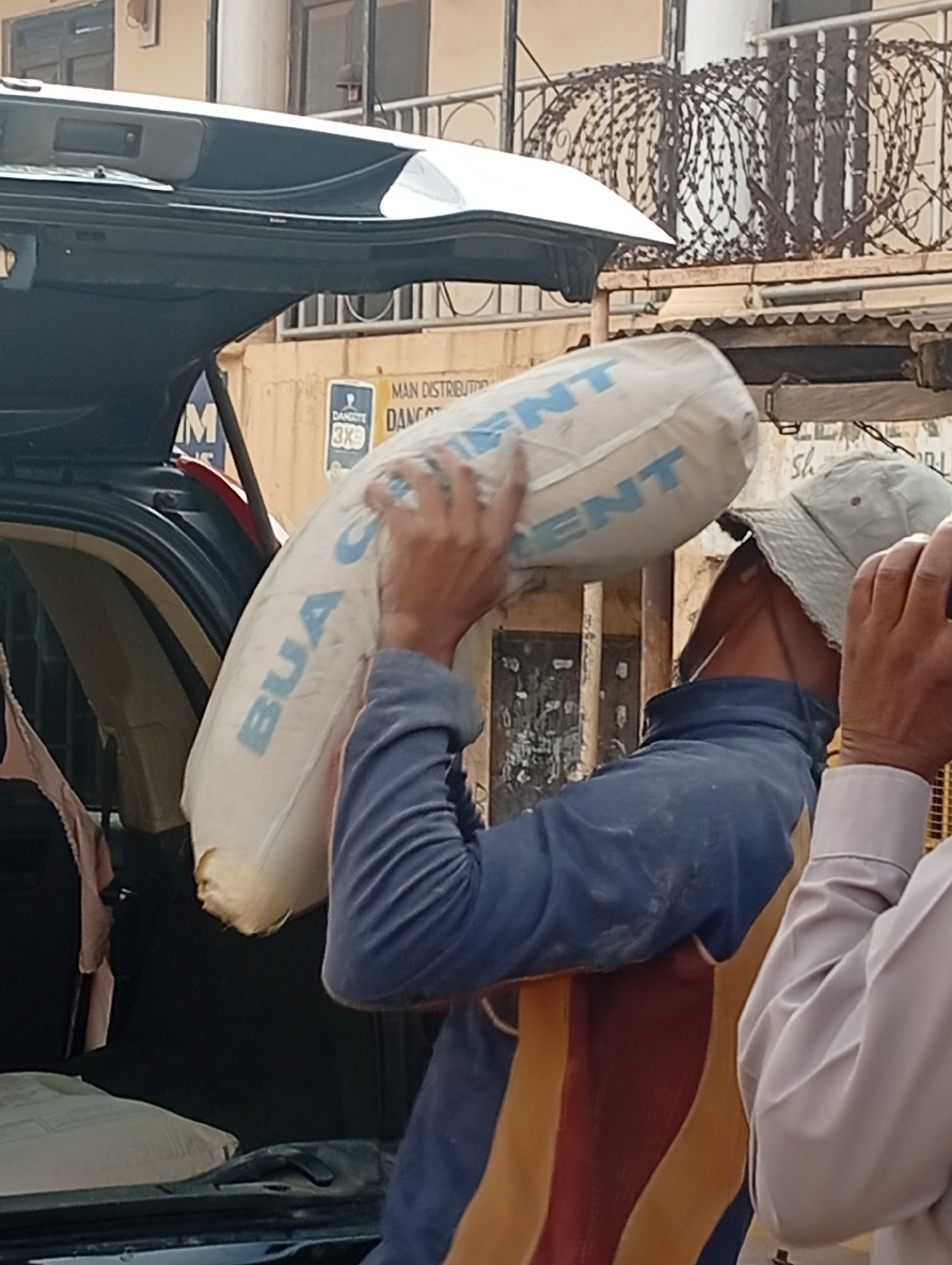 A man is carrying a bag of BUA Cement, presumably loading or unloading it from the open trunk of a car. The scene appears to be in the city of Gura Top, Nigeria, with a building in the background that has razor wire on the roof and signs for Dangote Cement, a main distributor, visible. Another man is partially visible to the right of the person carrying the cement bag.
