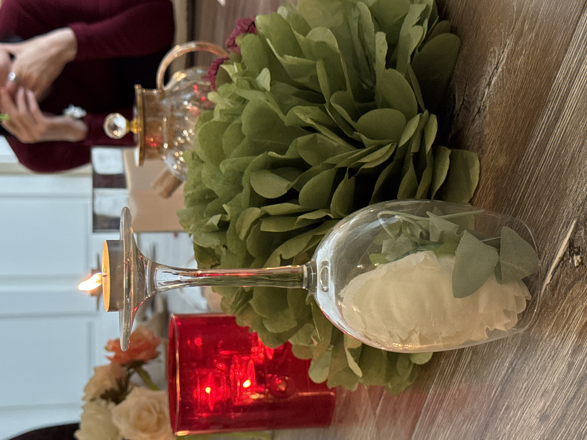 The image depicts a table setting, likely indoors, with a rustic wooden surface. At the forefront, a wine glass filled with a white flower and green foliage lies horizontally. Beside it, there is a green ornamental pom-pom. A candle holder, shaped like a tall glass with a flat top, holds a burning candle. A red glass container is also visible. In the background, a glass teapot and a person wearing a maroon long-sleeved shirt are partially visible. The location is Unknown, Unknown.