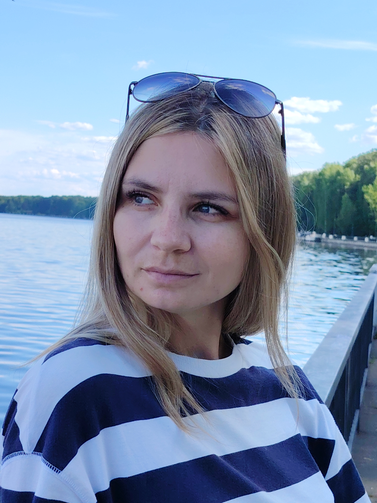 A woman is depicted outdoors next to a large body of water in Chaikovsky, Russia. She has light-colored, long hair and blue eyes, and is looking towards the left side of the frame with a neutral expression. Aviator-style sunglasses with reflective dark lenses are perched on top of her head. She is wearing a long-sleeved shirt featuring broad horizontal dark blue and white stripes. The background shows a calm blue body of water with subtle ripples, a distant treeline composed of dense green foliage, and a clear blue sky with scattered white clouds. A dark grey railing is visible along the right edge of the frame, indicating a pier or embankment.