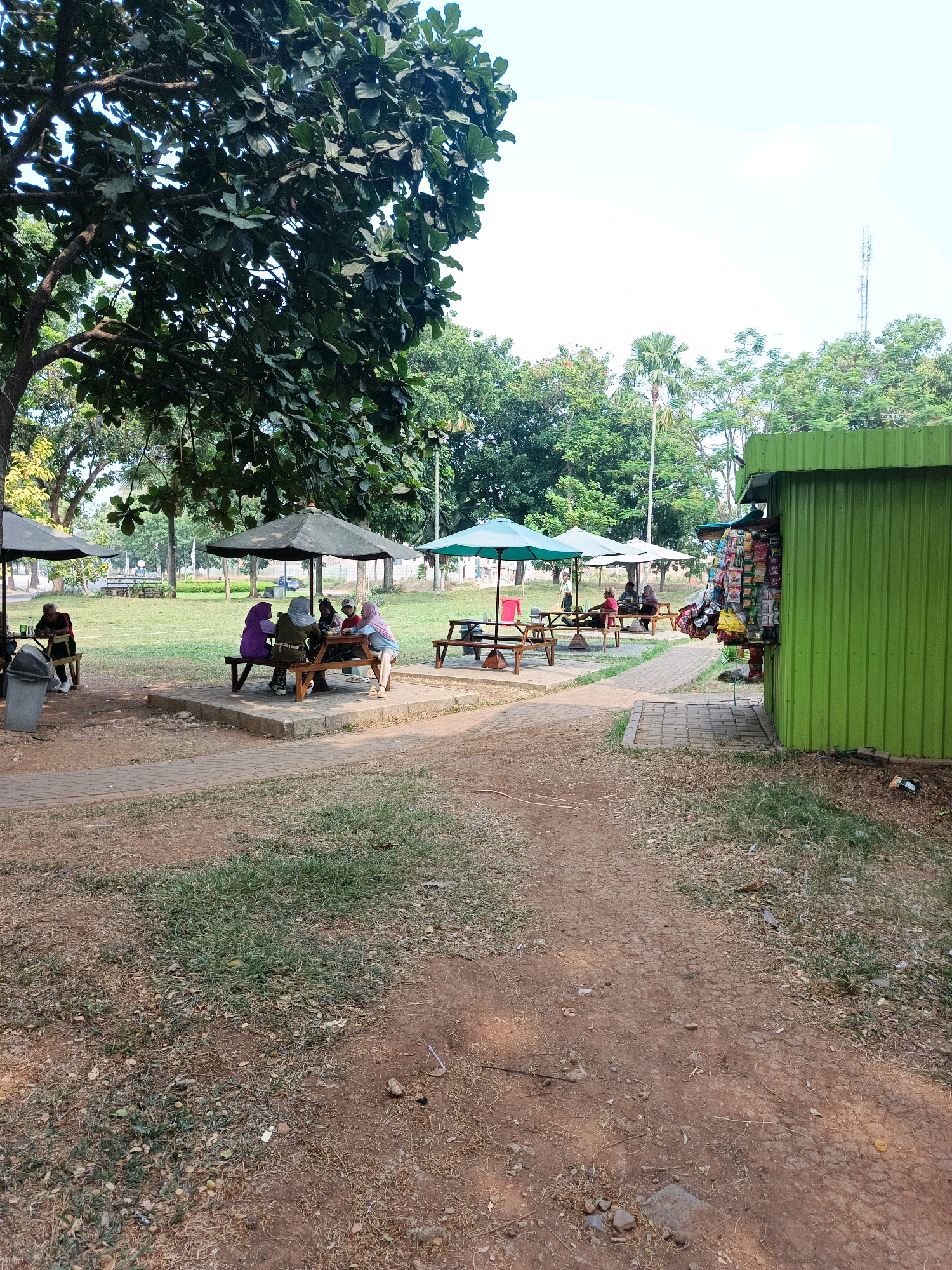 The image depicts an outdoor seating area in Sukamantri, Indonesia, likely a park or recreational space. Several people are seated at wooden picnic tables under umbrellas. Some tables are occupied by individuals or small groups, suggesting casual dining or socializing. To the right, a bright green corrugated metal kiosk is visible, stocked with various packaged goods displayed on its exterior, indicating it serves as a small shop or vendor. The ground is a mix of dirt paths and grassy areas, with trees providing shade. The overall environment appears to be a sunny day in a well-maintained public space.