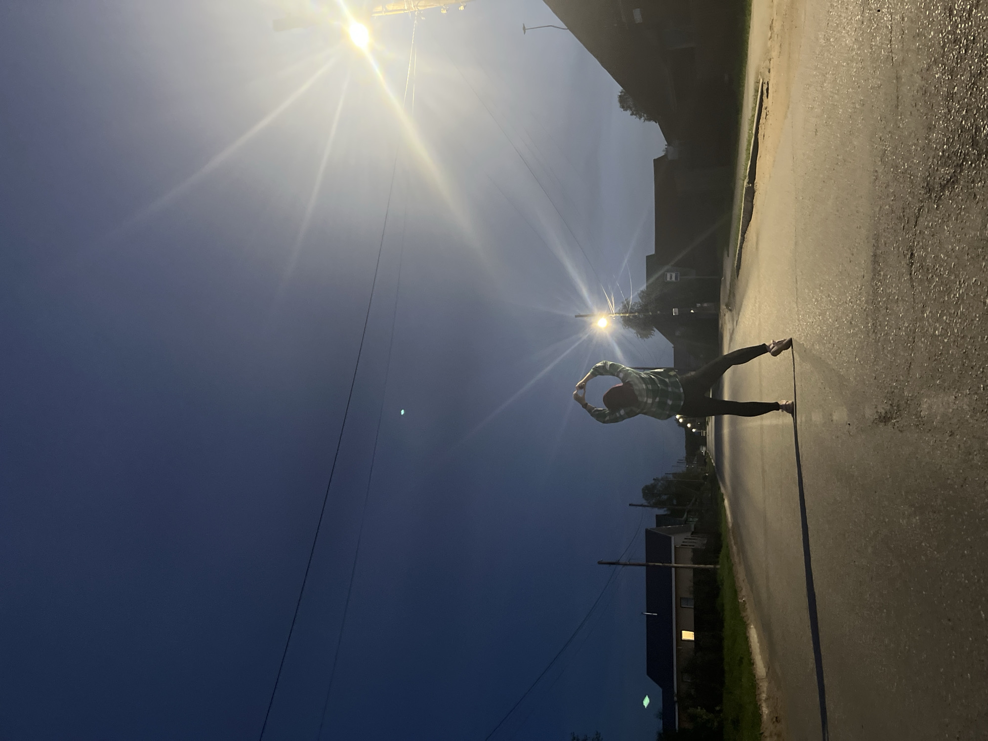 A person stands on a road under a streetlight, their arms forming a circle above their head. The sky is dark blue, and the road is wet. The location is Chaikovsky, Russia. Houses and trees line the side of the road.
