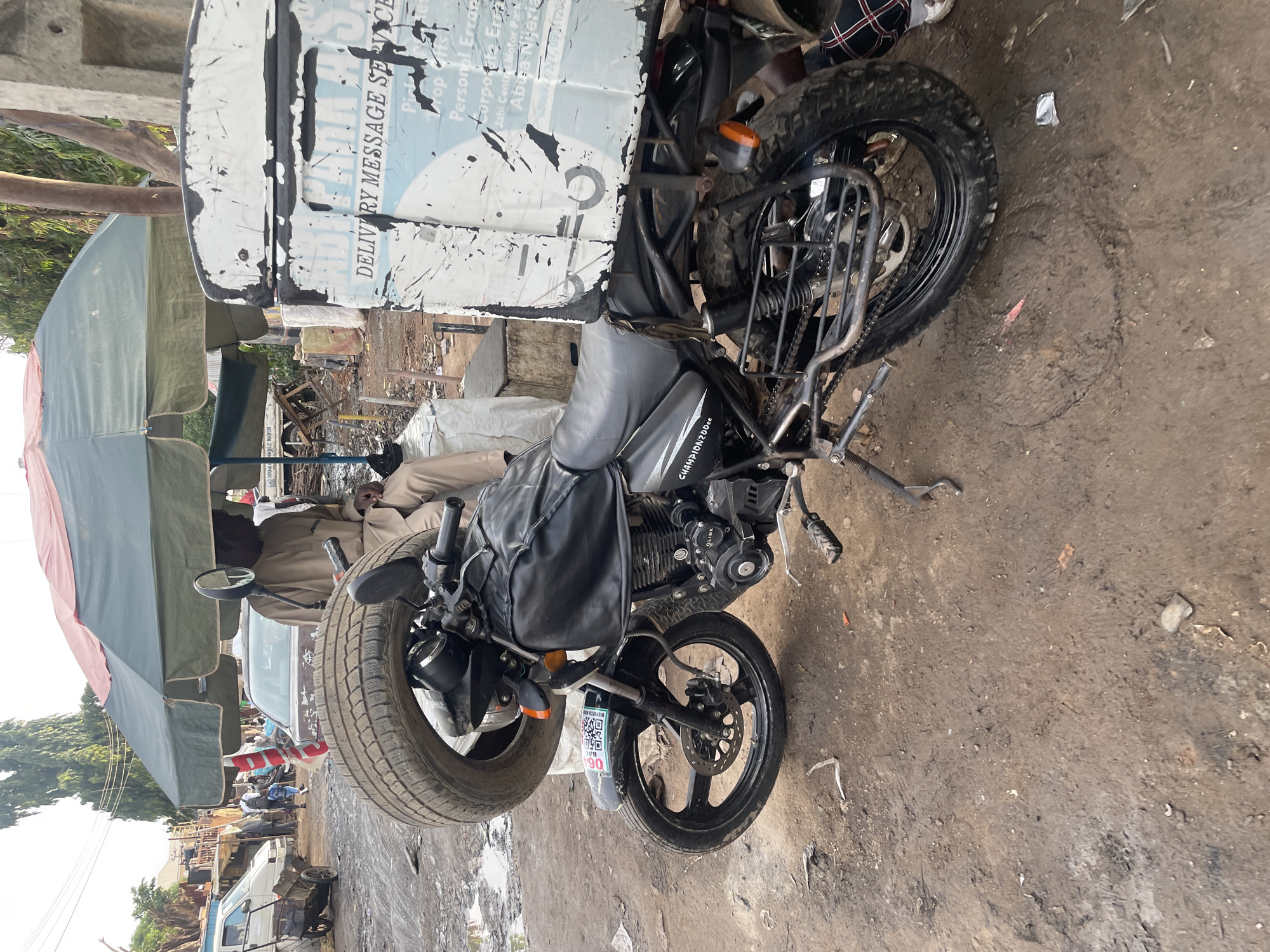 The image captures a black utility motorcycle parked outdoors in what appears to be a busy, unpaved area in Abuja, Nigeria. The motorcycle is equipped with black bags on its tank and an engine housing displaying "CHAMPION 200cc." A small plate near the front wheel shows a QR code and the number "064."
Attached to the rear of the motorcycle is a heavily worn, white delivery box with significant black scuff marks. Prominently visible text on this box reads "DELIVERY MESSAGE SERVICE," indicating its purpose. Further details on the box include service offerings like "Pick up," "Drop off," "Personal Errands," and "Corporate Errands." Location cues are also printed: "Jabi Centre Motor Park" and "Abuja Nigeria," along with phone numbers "6866920227" and "09011208." The text "OLINK" and "DISPATCH" are also partially visible, likely related to the delivery service.
The ground beneath and around the motorcycle is muddy and unpaved. In the background, there are several large green and light-colored market umbrellas, suggesting a street market or vendor area. Partial views of people are visible, including one person in a light-colored outfit seated under an umbrella and another with a plaid shirt near the motorcycle's rear wheel. Other vehicles, including a white van, are discernible in the distance. The scene is brightly lit, indicating daytime. The overall impression is one of a functional vehicle operating in a working urban environment.