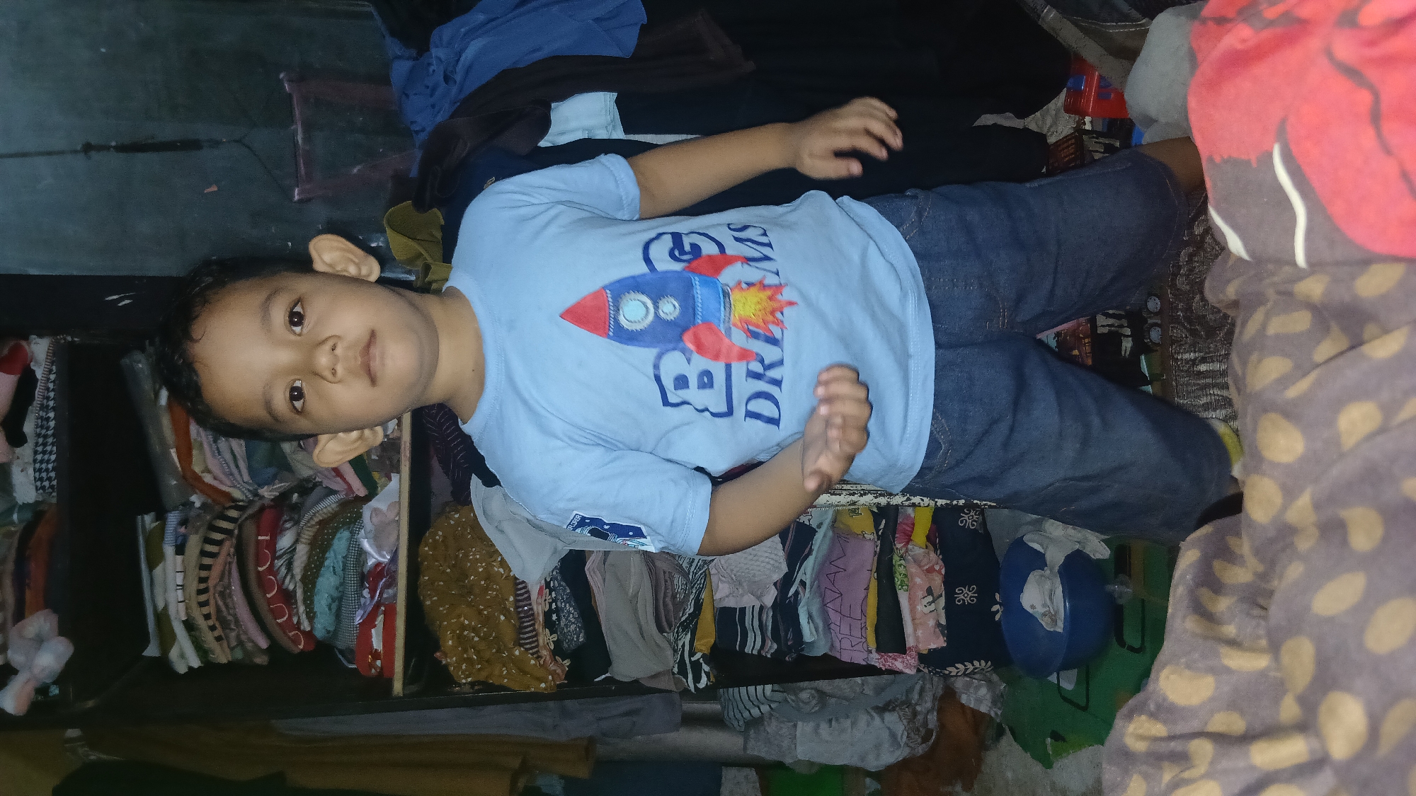 A young boy, appearing to be of Southeast Asian descent, stands in a cluttered room. He has dark hair and large ears. He is wearing a light blue t-shirt with a rocket graphic and the words "BIG DREAMS" printed on it, along with blue denim shorts. His left hand is held out, palm up, and his right hand is by his side. Behind him, a tall, dark shelving unit is overflowing with neatly folded clothes. To his right, a dark blue garment is draped over something out of frame. In the foreground, a bed or sofa covered with a brown and yellow polka dot patterned blanket is visible. The overall environment suggests a residential setting, possibly within a home in Keudejungkagajah, Indonesia, characterized by the presence of personal belongings and storage of clothing.
