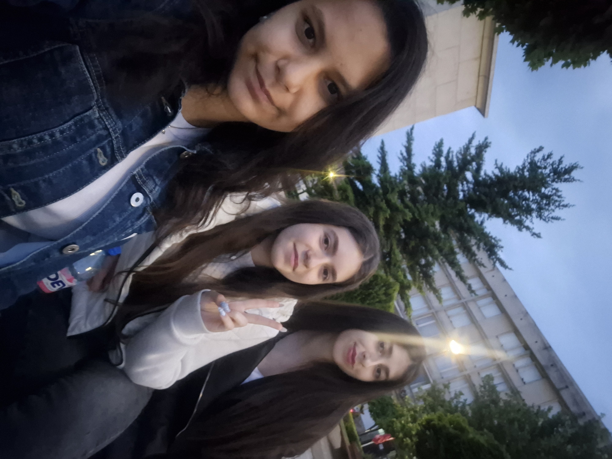 Three young women are posing for a selfie outdoors. The background includes a building and greenery, likely trees or bushes. The photo seems to have been taken in the evening or late afternoon, judging by the light in the sky. The middle woman holds up the 'peace' or 'victory' sign with her fingers. A water bottle is visible on the left side of the frame. The setting is likely a park or other public area in an urban environment in Pavlikeni, Bulgaria, as indicated.