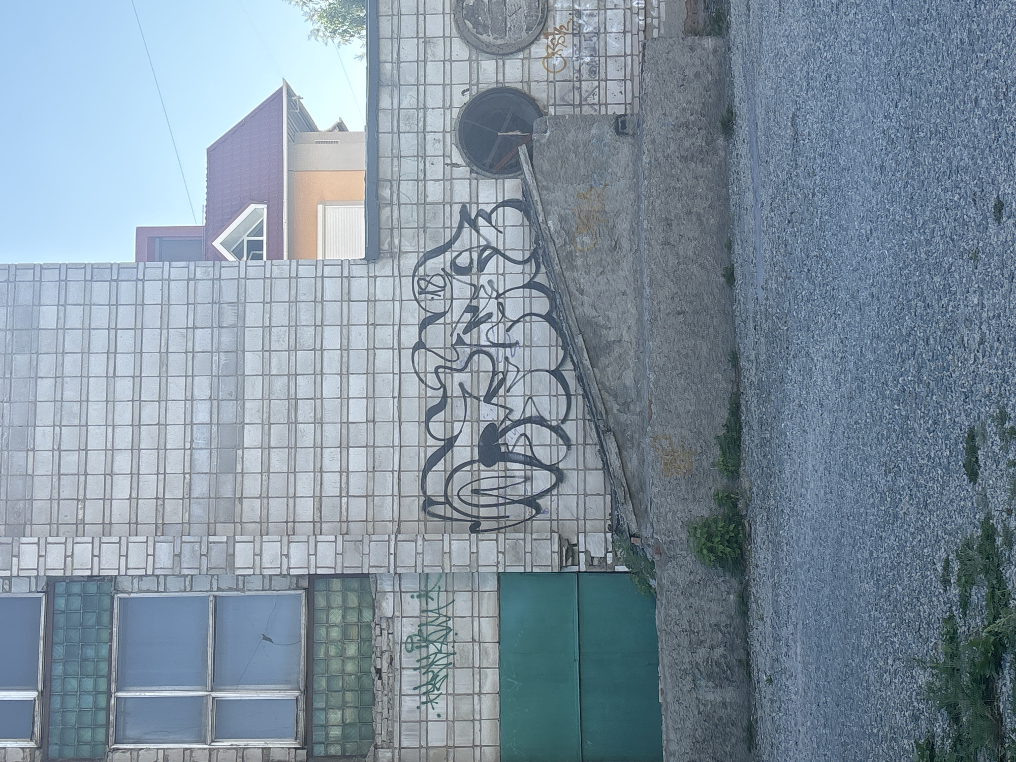 This is a medium-shot, eye-level, outdoor photograph taken during the day in Novosibirsk, Russia. The scene depicts the exterior of a multi-story building with a white tiled facade. The building features a large green metal door on the left side, and several windows, some of which are made of green glass blocks.
Graffiti is visible on the wall, including a large, black, stylized tag on the right side. To the left of this, a smaller green tag is present. In the background, a portion of another building with a red roof and yellow siding is visible. The ground in front of the building is covered with gravel.
There are no people or animals present in the image. The environment appears to be urban, with the building suggesting a commercial or industrial setting. The sky is clear and blue.