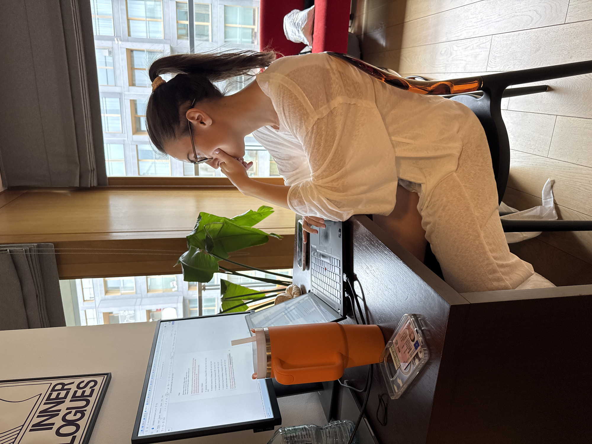 The image depicts a woman with her hair tied up in a ponytail, wearing glasses and a light-colored, loose-fitting top and possibly pants. She is seated at a desk, resting her chin on her hand, seemingly focused on the laptop and monitor in front of her. The laptop is open, and the monitor displays text. On the desk, there is a large orange tumbler with a straw, a cell phone in a clear case next to a license and some other items. A green leafy plant is also visible on the desk.
Behind the woman and visible through the window are tall buildings, suggesting an urban location. There are gray blinds covering the upper portion of the window. A red chair is partially visible in the background. The floor appears to be made of wood. A framed piece of art with the words "INNER LOGUES" is also visible.
