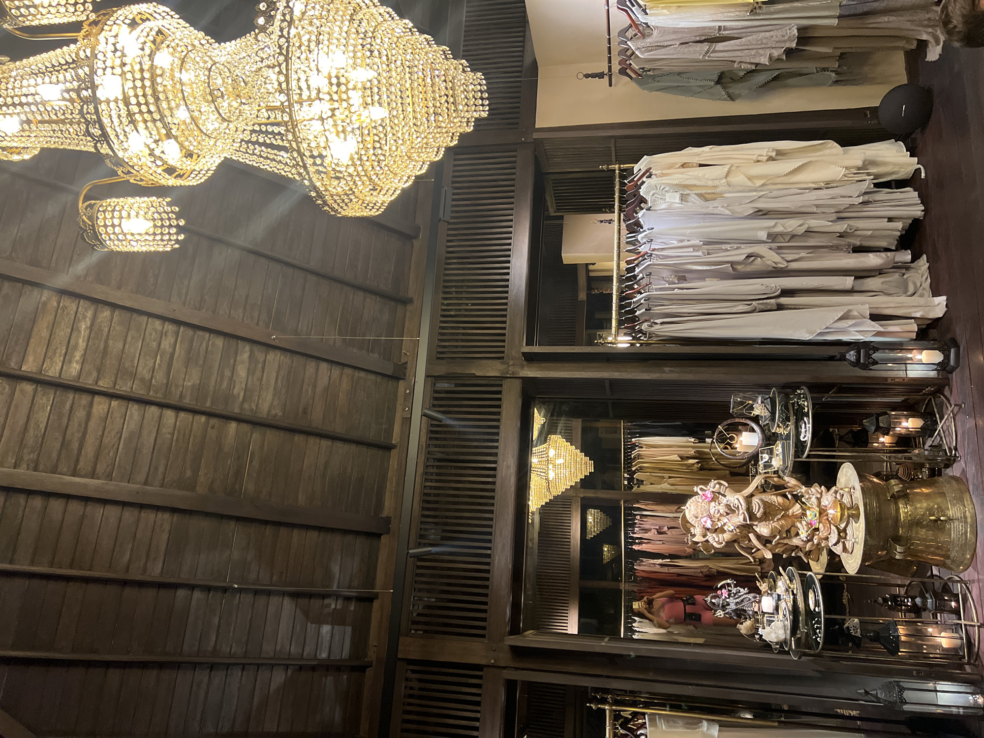 Captured in Ubud District, Indonesia, the scene depicts an interior space with a high, wooden-paneled ceiling from which hangs a large, multi-tiered chandelier. To the right, there is a closet with racks of clothing. The lower shelves display decorative items and sculptures. The image is taken from a low angle, emphasizing the verticality of the room and the grandeur of the chandelier. The space exhibits a combination of rustic and luxurious elements.