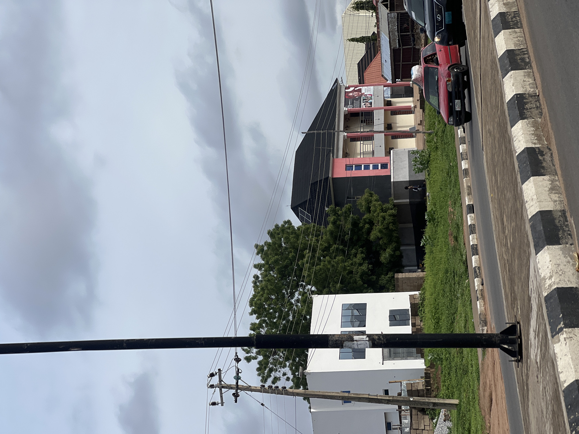 This image depicts a street scene in Jalingo, Nigeria, under a cloudy sky. A black pole stands in the foreground, positioned on the right side of the frame, its base secured with metal bolts. To the left of the pole, a wooden utility pole with ceramic insulators and power lines is visible. Behind the utility pole, a modern, white, two-story building with large dark-paned windows occupies the left portion of the background.
Further into the background, a vibrant pink and gray building with multiple balconies and a black pitched roof is prominent. A banner featuring faces and text is attached to this building. Adjacent to it, smaller structures with corrugated metal roofs are present. Green foliage, including a large tree, surrounds the buildings and lines the street.
The street itself features a paved road with a distinct black and white striped curb. Two vehicles are visible on the road: a red hatchback in the center of the frame, with a person visible in the driver's seat and what appears to be a white object secured to its roof, and a dark-colored SUV to the right, partially obscuring the red car. A narrow patch of green grass and some construction materials are present between the road and the buildings.