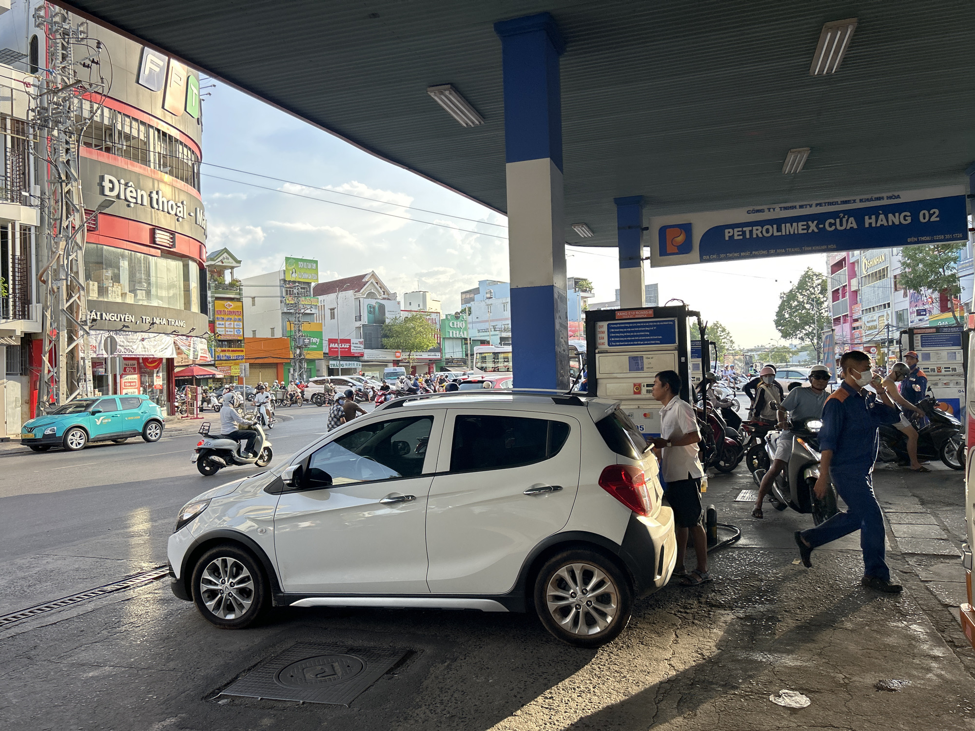 A sunny day in Tay Nha Trang Ward, Vietnam, depicts a busy street scene at a PETROLIMEX gas station. The foreground features a white car parked at a pump, with a person standing next to it. To the left, a teal car is visible on the road, and numerous motorcycles and scooters navigate the street, some with riders. In the background, buildings with signage in Vietnamese line the street, indicating shops and businesses. The gas station itself has a blue and white striped roof structure supported by pillars, with various gas pumps and informational signs. Several individuals, some in work uniforms and others in casual attire, are present at the gas station, interacting with vehicles and equipment. The overall atmosphere suggests a typical urban environment with ongoing commercial activity.