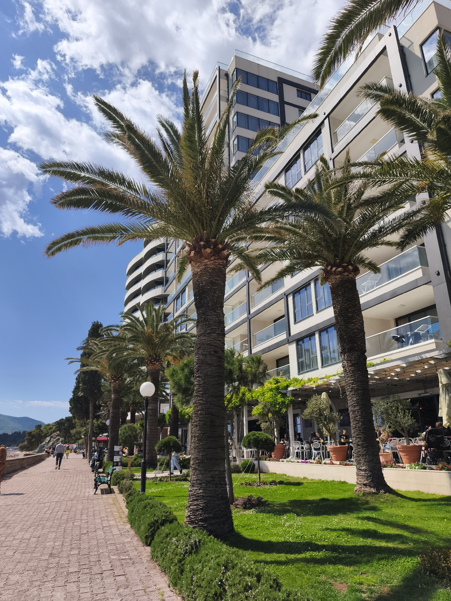 The image captures a sunny scene in Bečići, Montenegro, along a brick-paved walkway lined with palm trees and lush greenery. The walkway is occupied by a few pedestrians in casual attire. Adjacent to the walkway is a well-maintained grass area, where the shadows of the palm trees fall. Further in, a modern building with multiple balconies and large windows provides a backdrop. At ground level, a restaurant or cafe area is visible, with people seated at tables under a pergola covered in greenery. The sky above is blue with scattered white clouds.