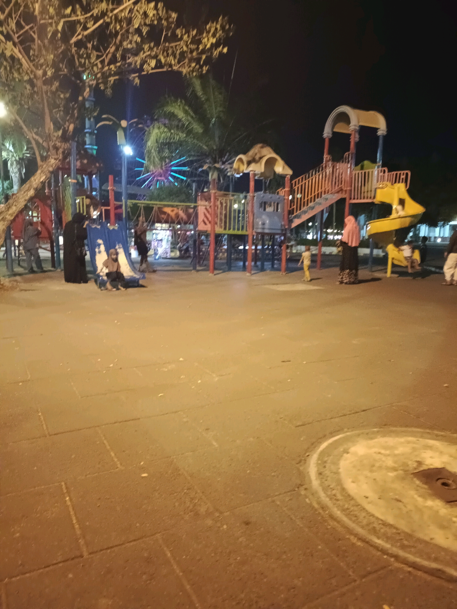 An outdoor public playground is illuminated by artificial lights at night. The playground features a colorful slide structure with red and yellow elements, and a blue slide. Several individuals are present; a woman in a black abaya and headscarf stands near the blue slide, while another woman in a pink headscarf and patterned skirt is by the yellow slide. A young child in a yellow shirt and pants stands near the pink-scarfed woman, and another child is on the yellow slide. In the background, a Ferris wheel with blue lights is visible, along with trees and palm trees. The ground is paved with square stones. The scene appears to be in Lamongan, Indonesia, a city known for its parks and recreational areas.