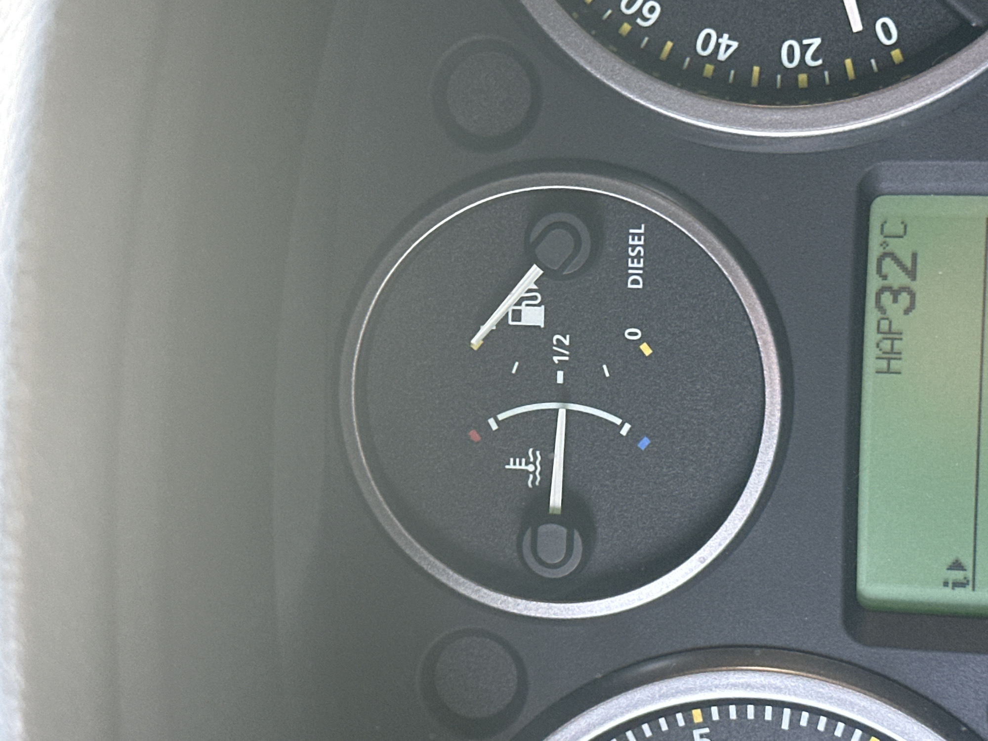 This media file shows a close-up of a vehicle's dashboard. The primary focus is a fuel gauge for a diesel engine, indicated by the text "DIESEL" to the right of the gauge. The needle on the fuel gauge is positioned slightly above the "1/2" mark, suggesting the fuel tank is more than half full. Below the fuel gauge is a temperature gauge with markings for cold (red) and hot (blue), and the needle is positioned in the middle range, indicating a normal operating temperature. To the right of these gauges is a digital display showing "HAP32°C", likely indicating an outside temperature of 32 degrees Celsius. The dashboard appears to be from an older vehicle, with a slightly worn texture. In the upper part of the image, a portion of another gauge, possibly a speedometer, is visible with markings up to 60. The lighting suggests it is daytime, and the overall scene is that of a car interior, likely in an urban or suburban setting given the reference to "Khimki, Russia" as a location context, though no specific external environment is visible. There are no people or animals present in the frame.